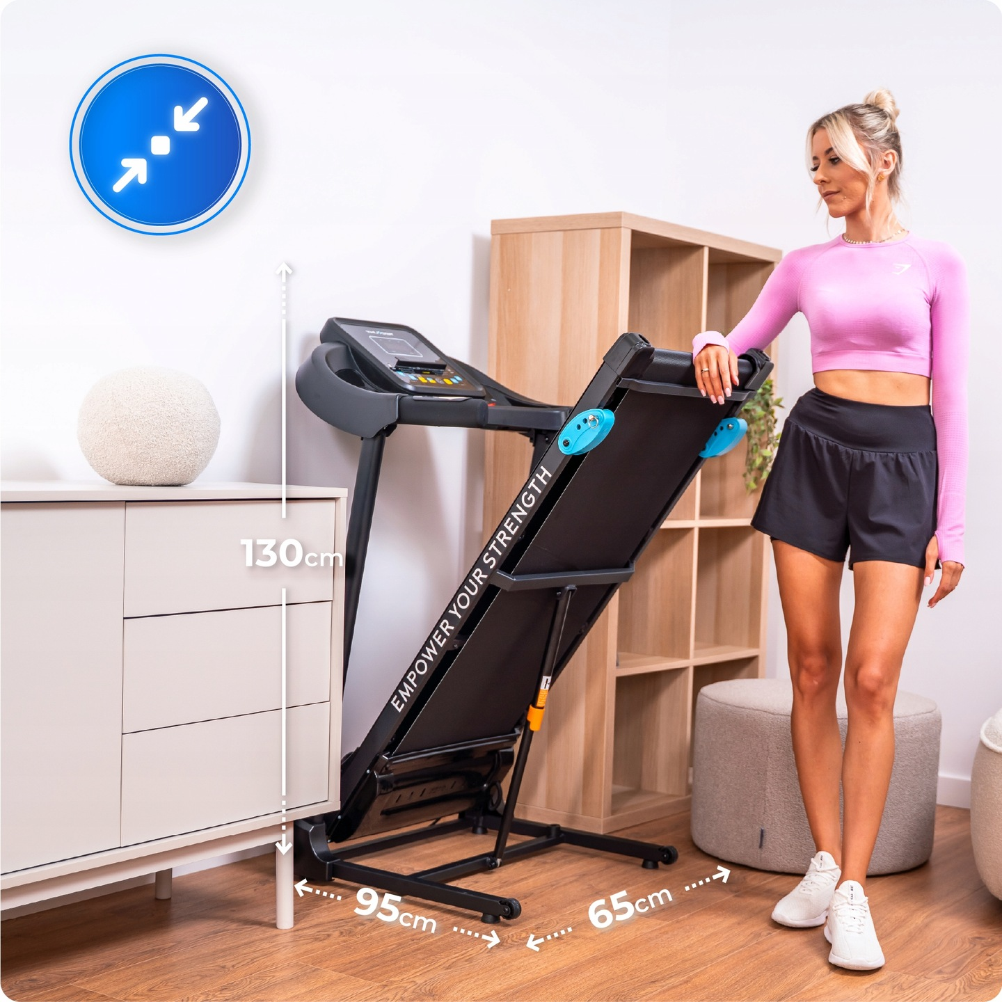 Kobieta w stroju sportowym z bieżnią. Białe buty, czarne szorty, różowy top. Pokój z meblami, oznaczone wymiary.