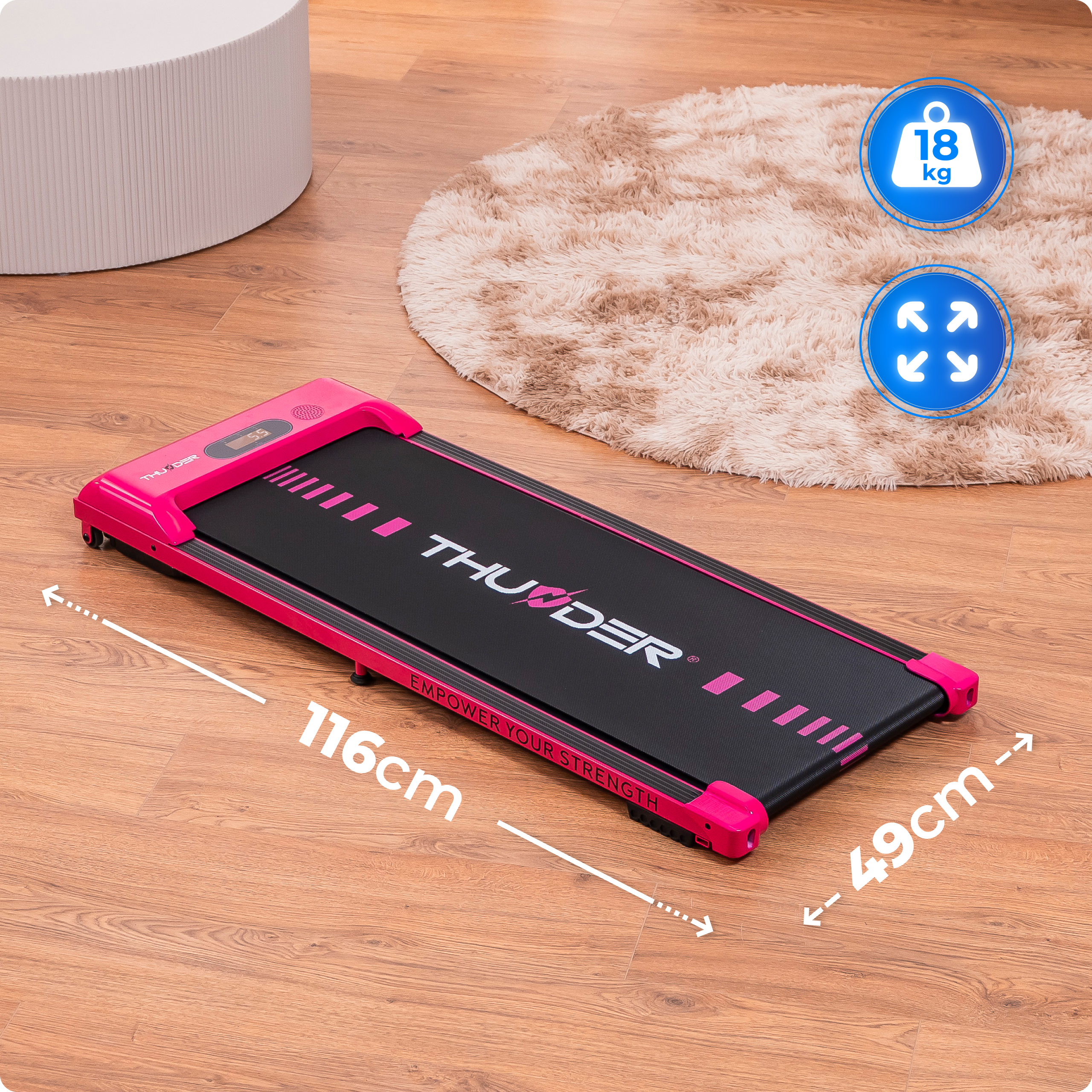 Różowa bieżnia na drewnianej podłodze, obok puszystego dywanu i białego walca. Pokazano wymiary. Wyświetlana waga 18 kg.