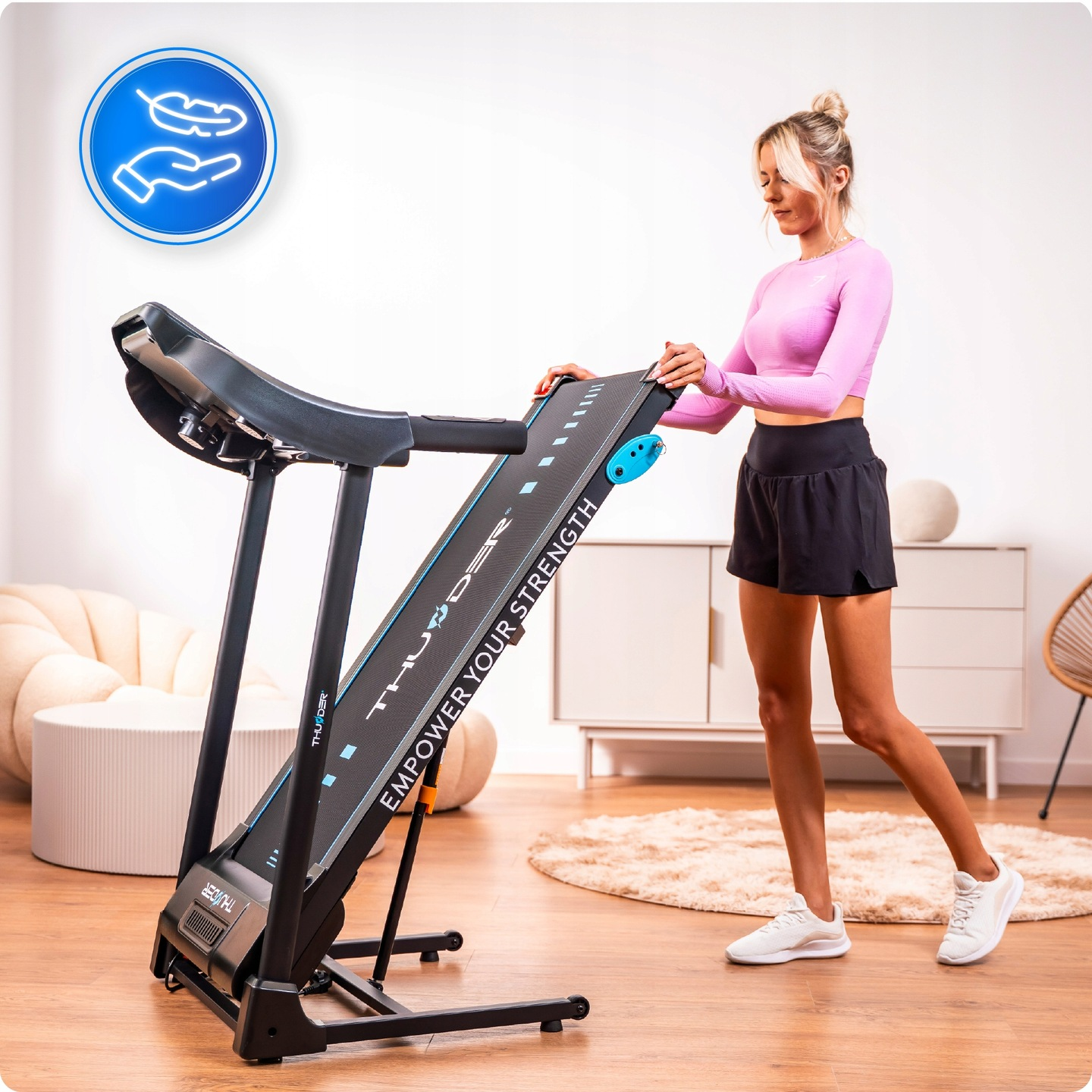 Kobieta obok bieżni. Ma na sobie różowy top i czarne szorty. Bieżnia jest czarno-niebieska, z nazwą marki.