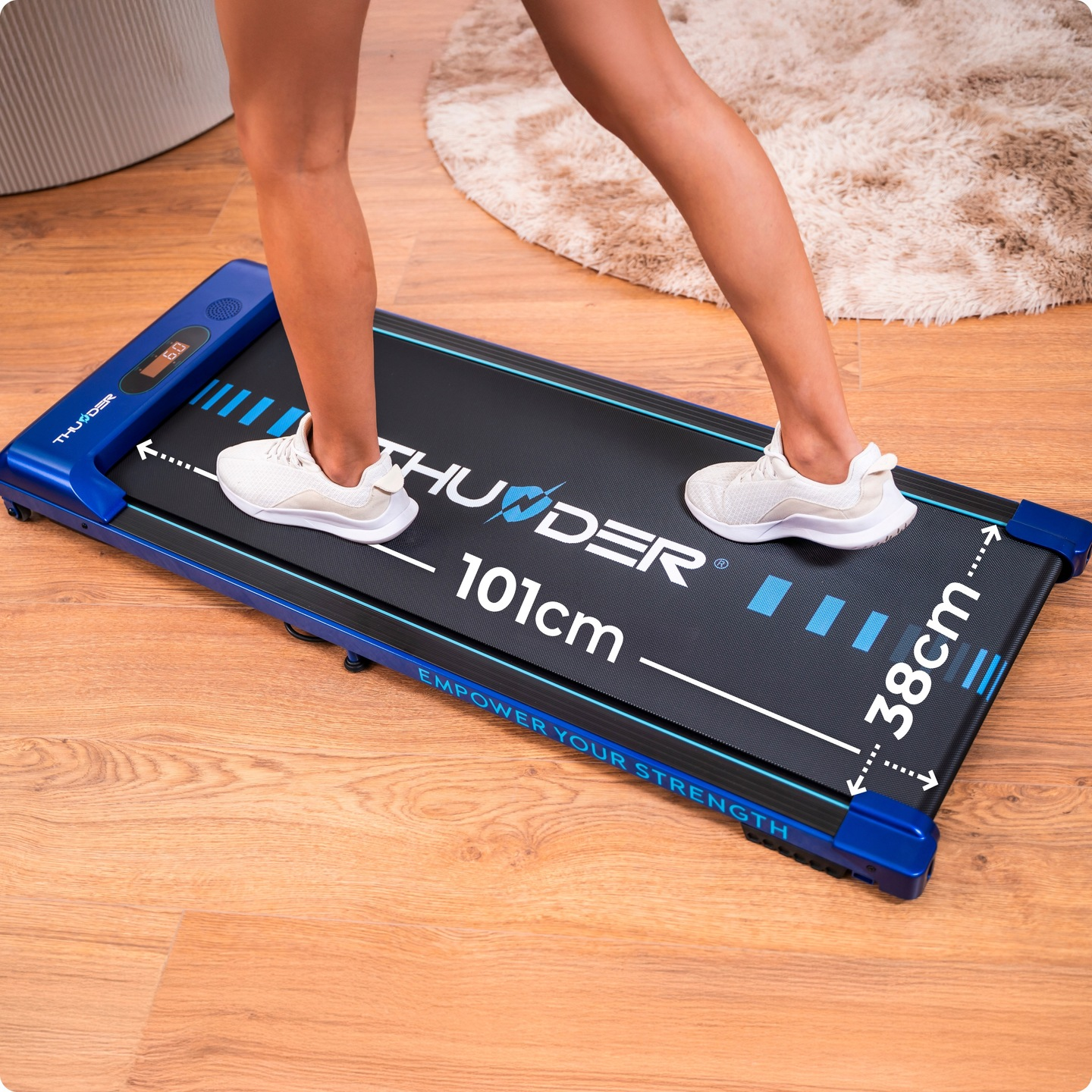 Osoba stoi na małej bieżni. Bieżnia jest czarno-niebieska i stoi na drewnianej podłodze. Osoba ma na sobie białe trampki.