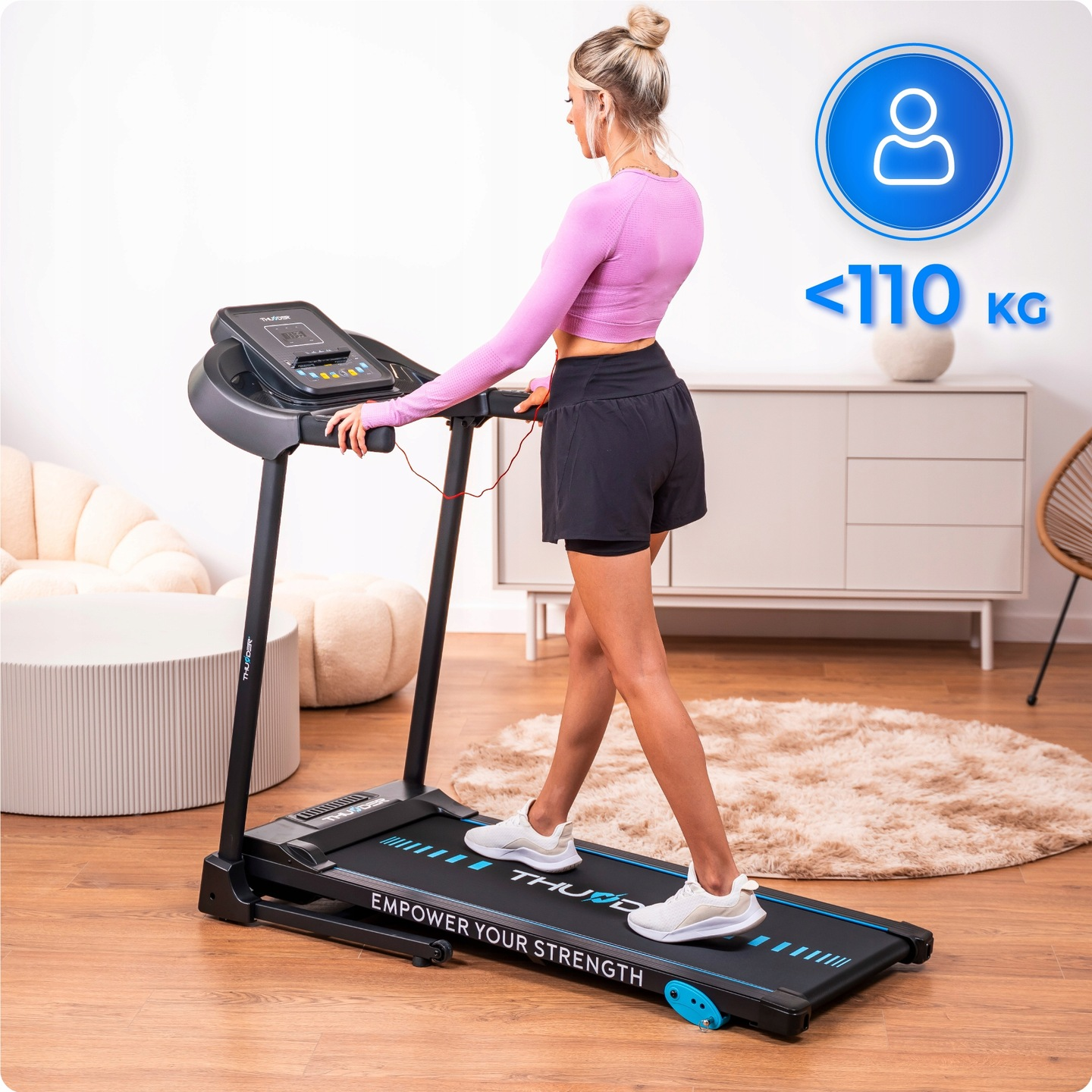 Kobieta ćwiczy na bieżni. Nosi strój sportowy i słuchawki. Bieżnia ma cyfrowy wyświetlacz. Widoczny jest znak ograniczenia wagi.