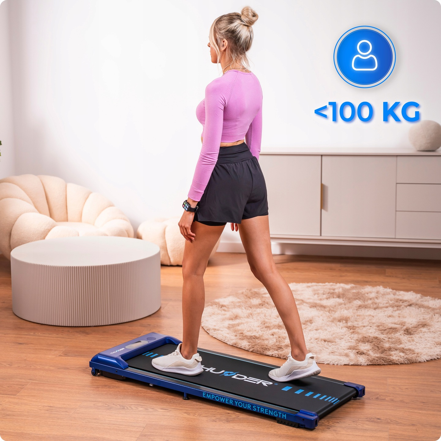 Kobieta w stroju sportowym ćwiczy na bieżni. Bieżnia jest niebiesko-czarna. W tle dywan i szafka.