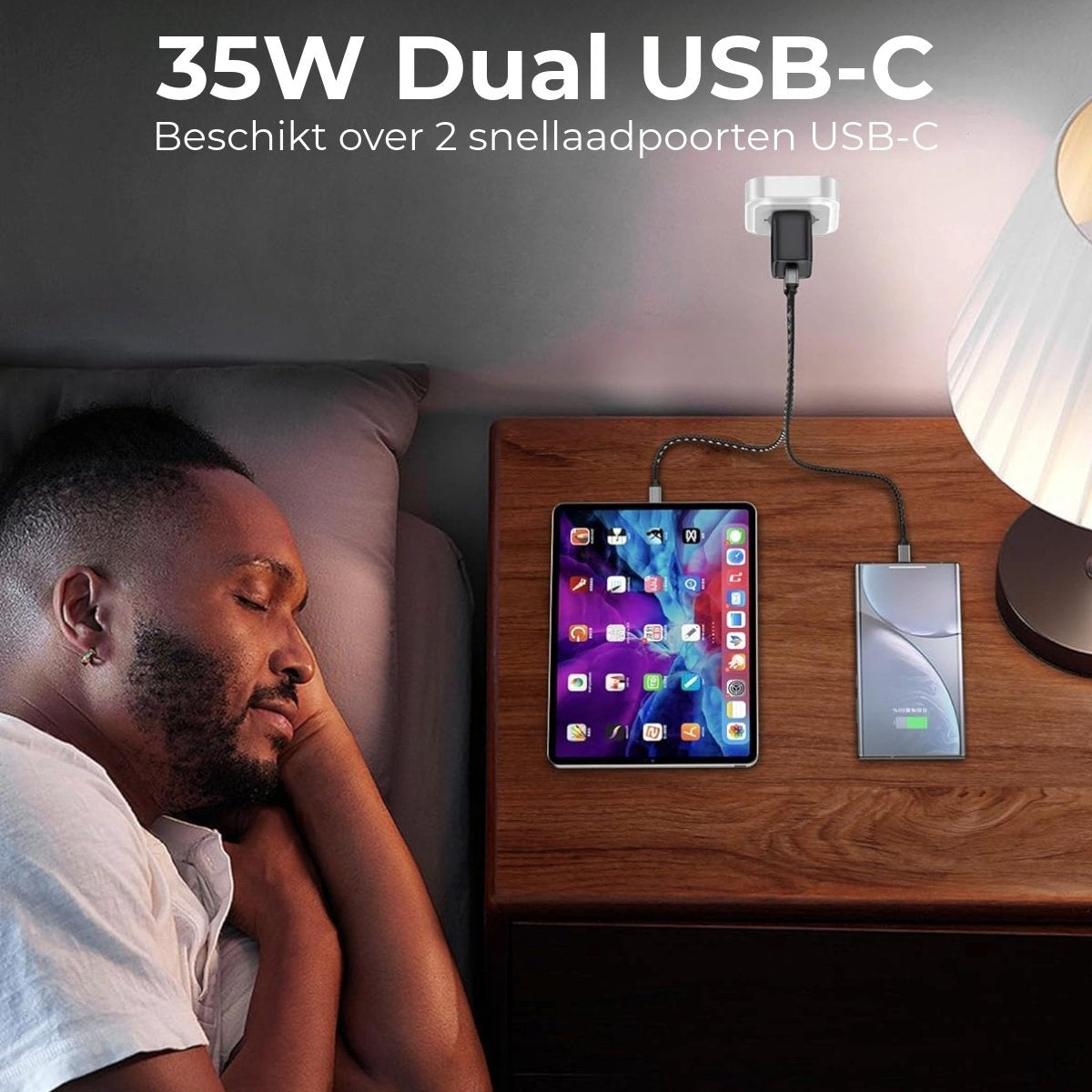 A man sleeps next to a nightstand with a tablet and a power bank charging. A lamp is next to them. USB-C charger on top.