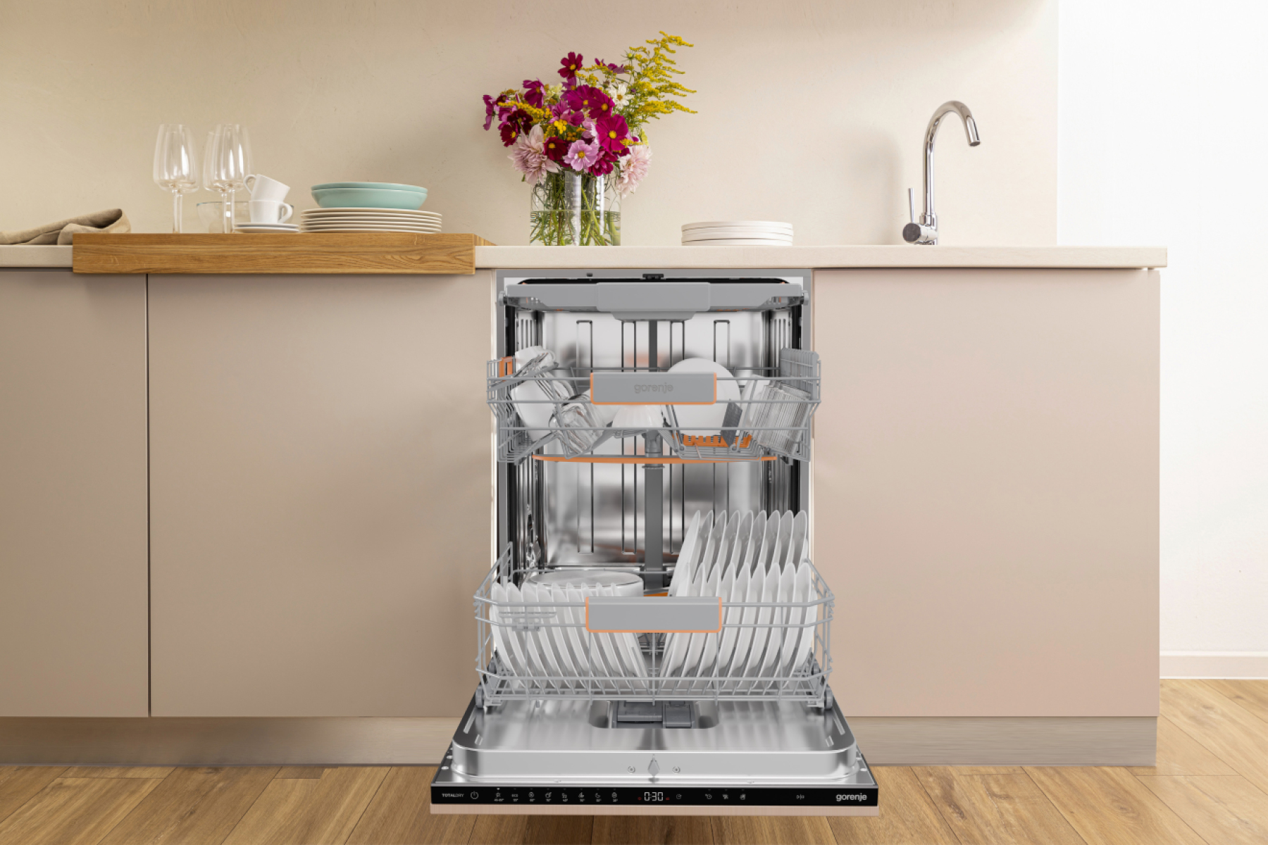 Otwarta zmywarka z naczyniami. Kwiaty i szklanki na blacie. Scena kuchenna z beżowymi szafkami i srebrną baterią.