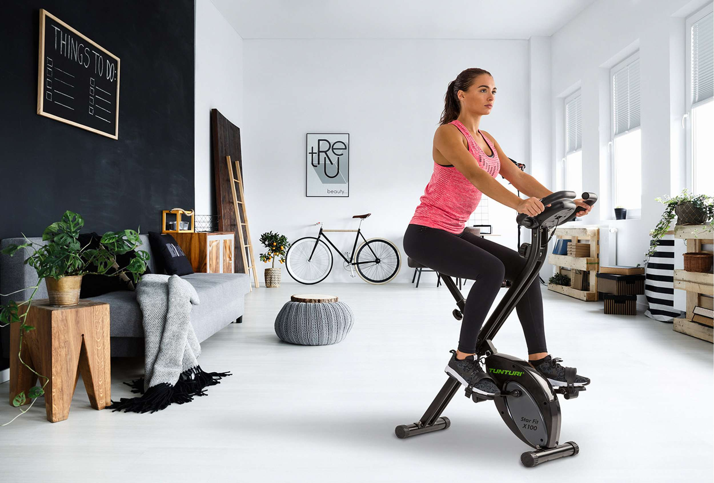 Vrouw traint op een hometrainer in een lichte kamer. Zwarte fiets en decoratie op de achtergrond.