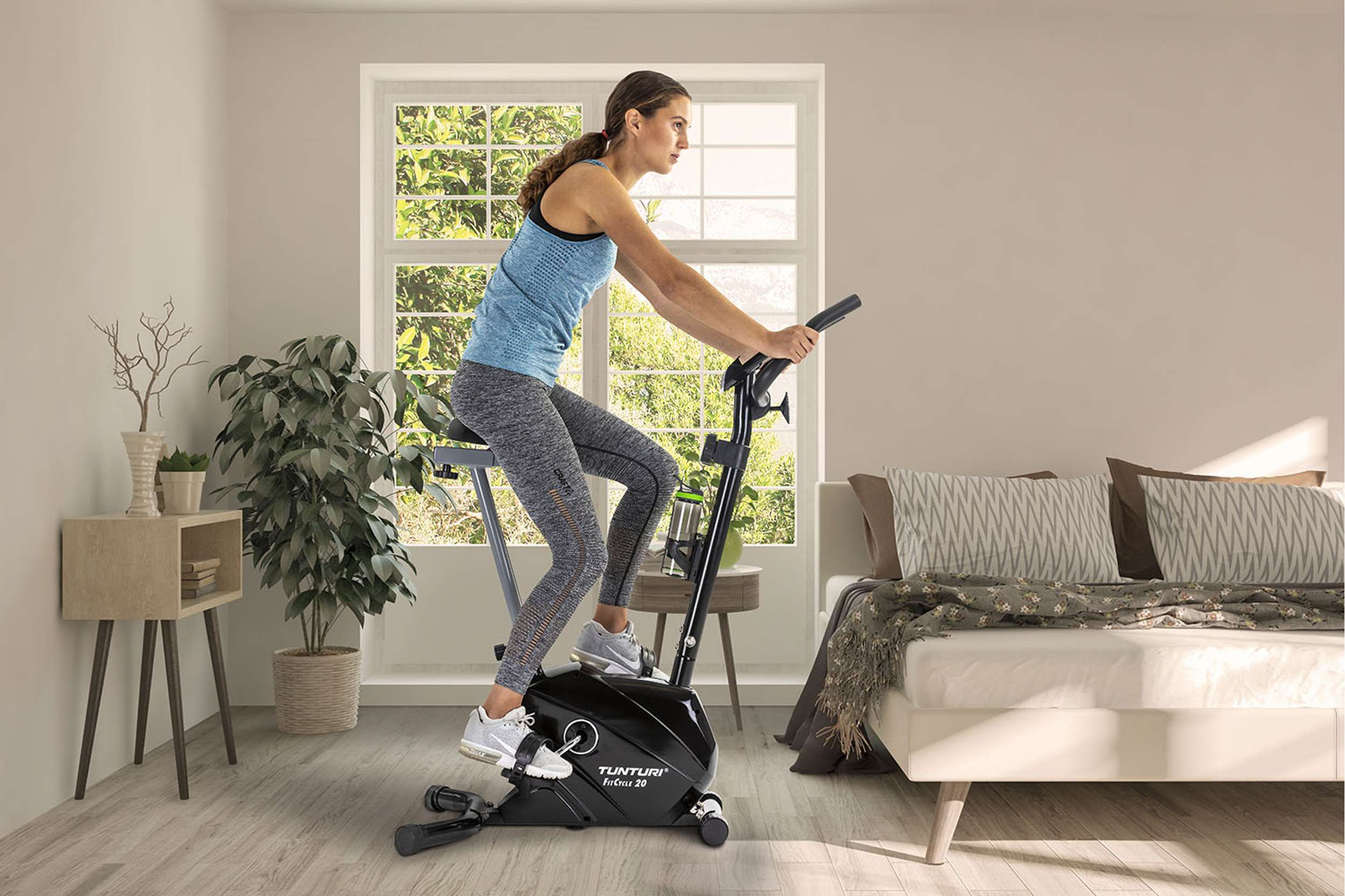Woman on exercise bike. She wears blue top, grey leggings, and sneakers. Room has bed, plants, and window.