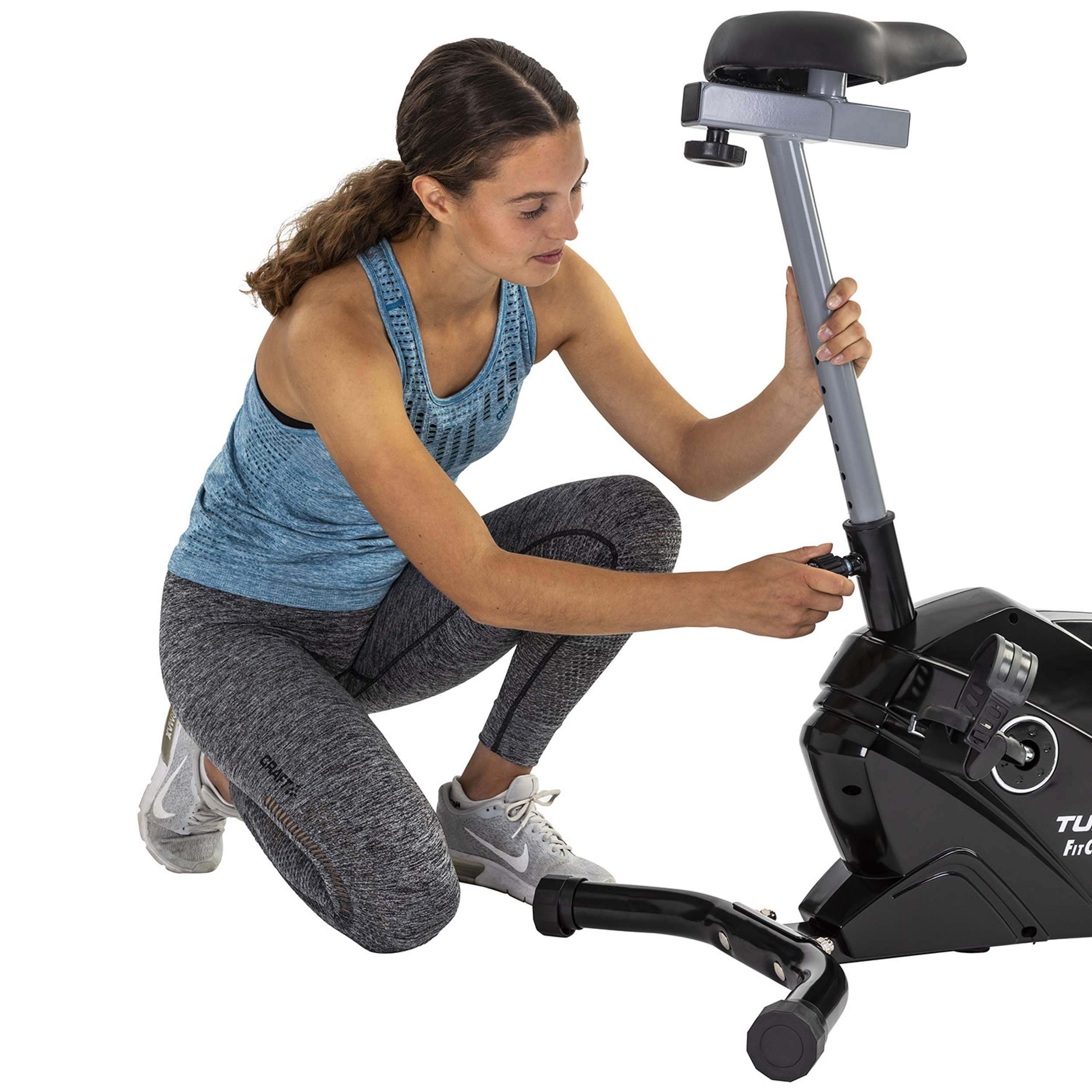 A woman assembles an exercise bike. She's wearing a blue tank top and gray leggings, squatting near the bike.