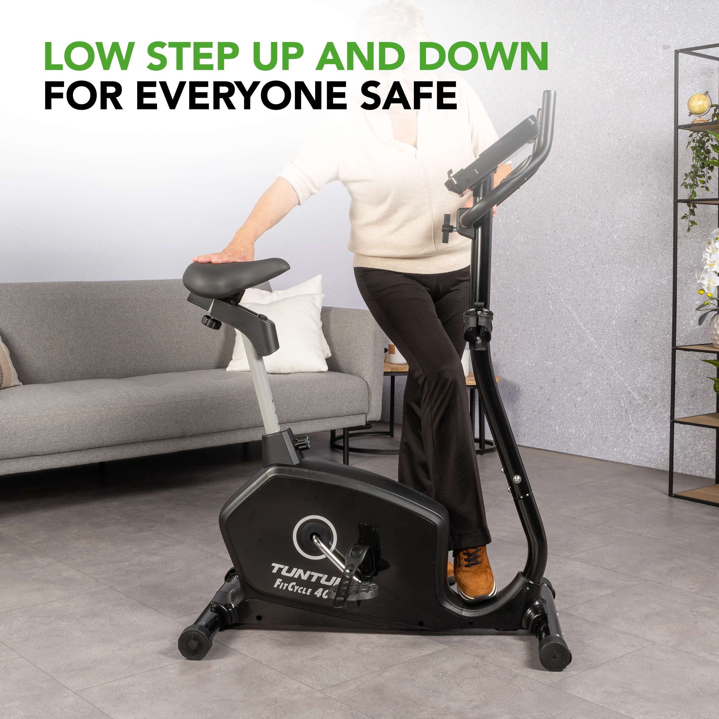Een oudere vrouw die een zwarte hometrainer gebruikt. De fiets staat in een kamer met een grijze bank en een plank.