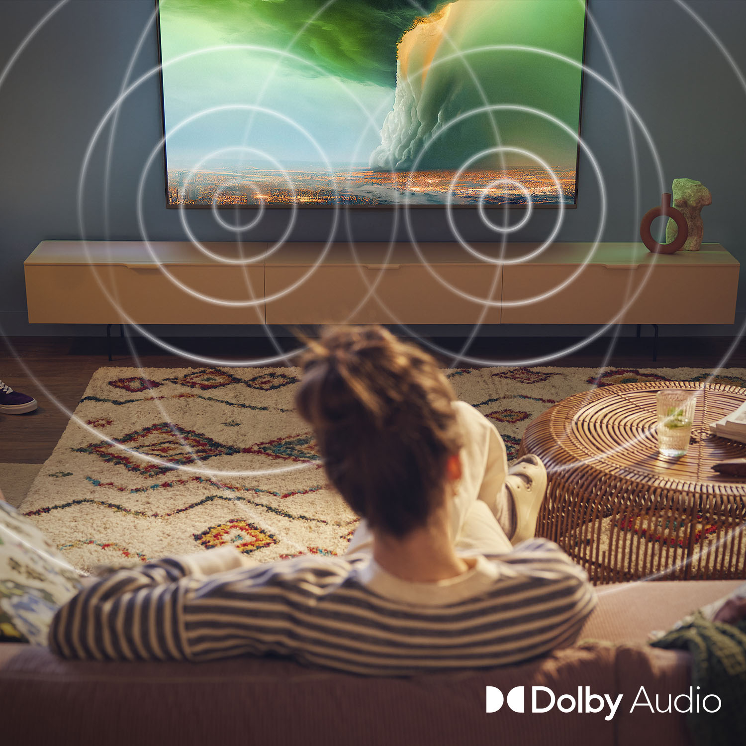 Osoba ogląda telewizję, z falami dźwiękowymi wychodzącymi z ekranu. W pokoju znajduje się dywan, kanapa i stolik kawowy.