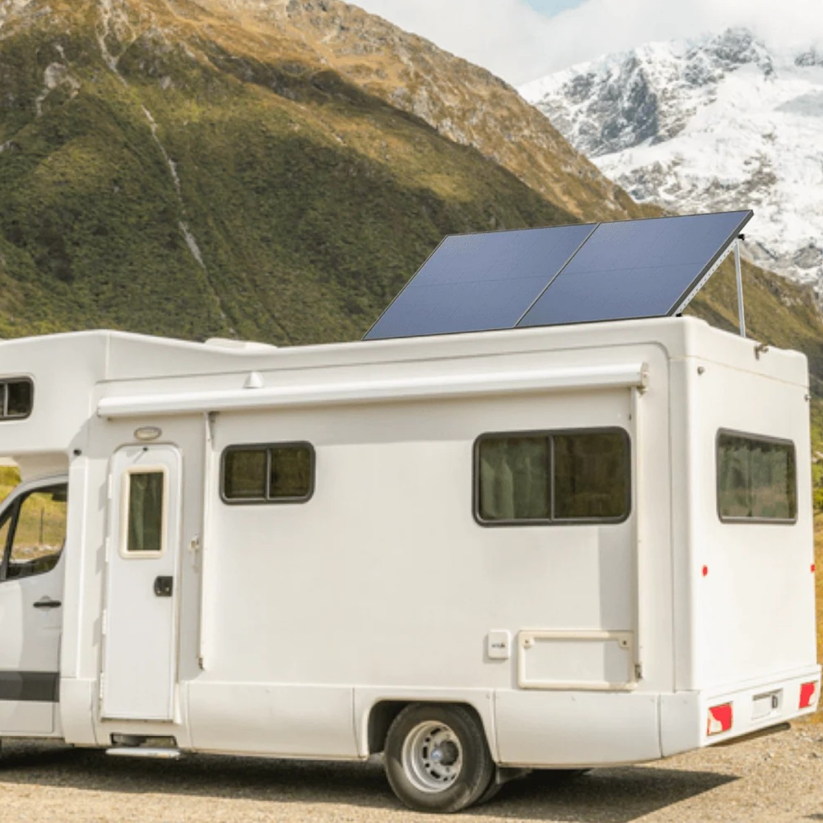 Camper bianco con pannelli solari sul tetto, parcheggiato davanti alle montagne. Colline verdi e cime innevate sullo sfondo.