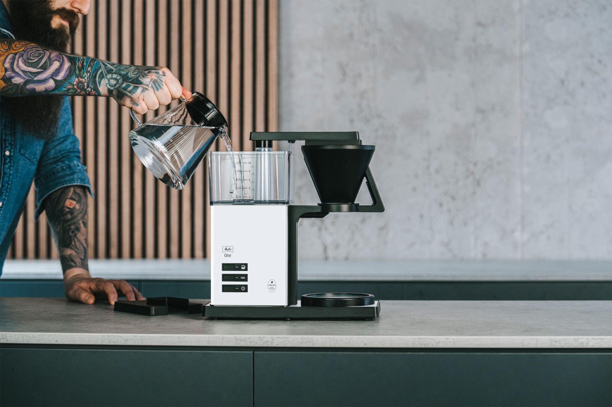 Ein Mann gießt Wasser aus einem Glaskrug in eine weiße Kaffeemaschine. Der Mann hat Tätowierungen. Der Hintergrund hat Holzpaneele und eine helle Wand.