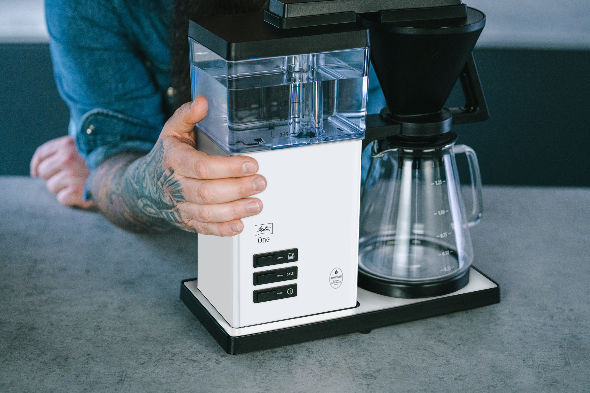 Eine tätowierte Person hält eine weiße Kaffeemaschine. Sie hat einen Wasserbehälter und einen schwarzen Filter. Eine Glaskaraffe ist sichtbar.
