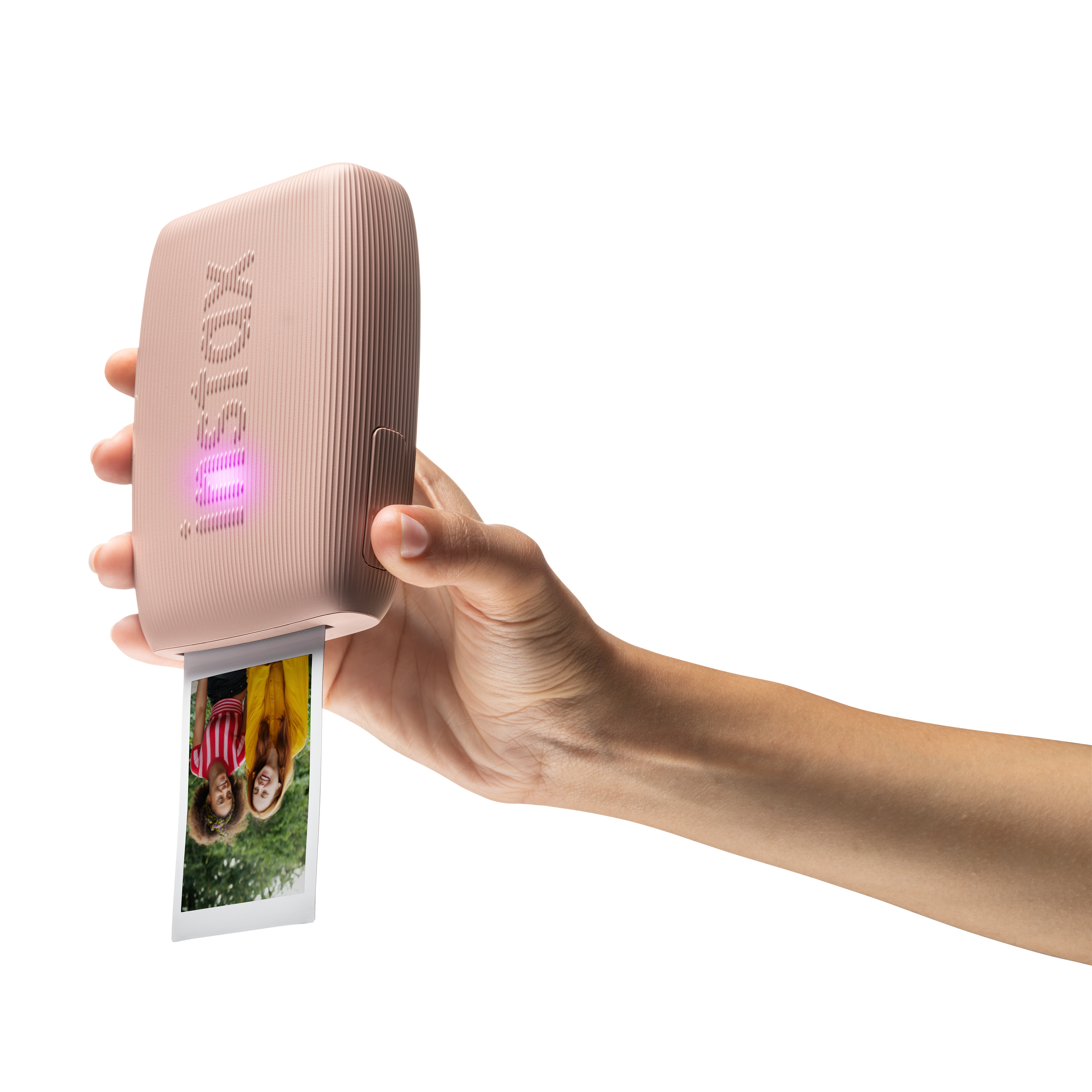 Una mano tiene una stampante Instax rosa, che stampa una foto di due donne sorridenti. Lo sfondo è bianco.