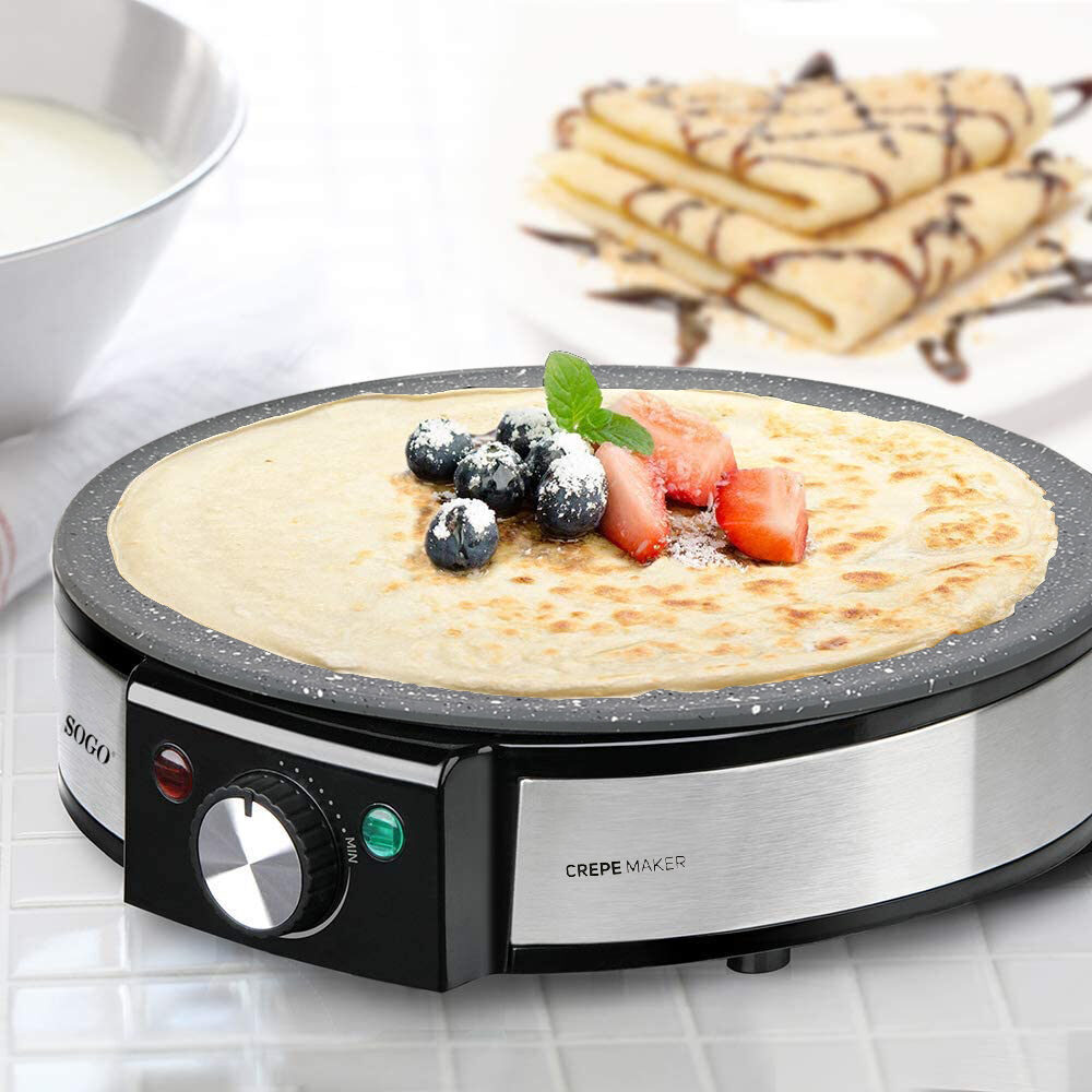 Crepera con un crepe cocido cubierto con arándanos y fresas. Plato de crepes al fondo. Un bol de leche a la izquierda.