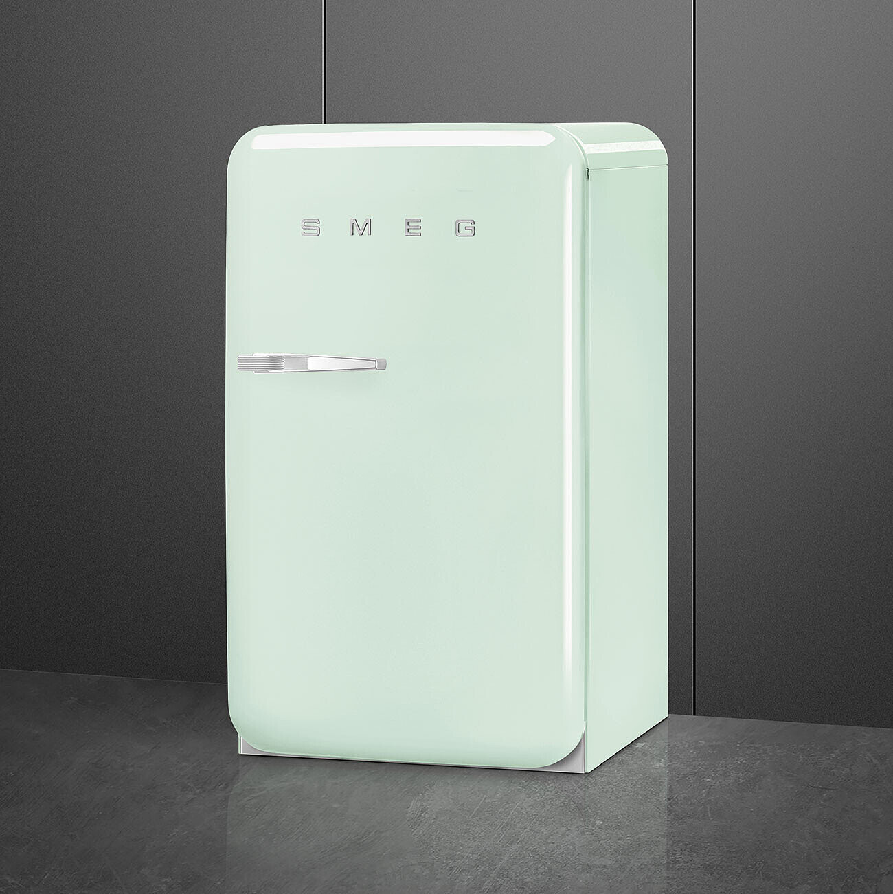 Un refrigerador SMEG verde menta de estilo retro sobre un suelo gris. El fondo es una pared gris oscura.