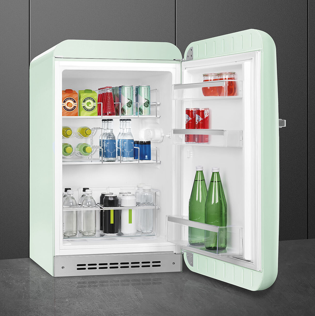 Mini-nevera retro abierta con bebidas. Contiene botellas, latas y cajas de zumo. Exterior verde menta, interior y estantes de acero inoxidable.
