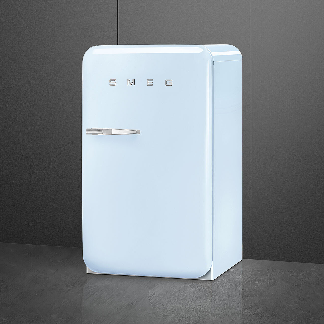 Un refrigerador SMEG celeste claro se encuentra frente a una pared y un piso grises oscuros. El mango es plateado y la marca es visible.