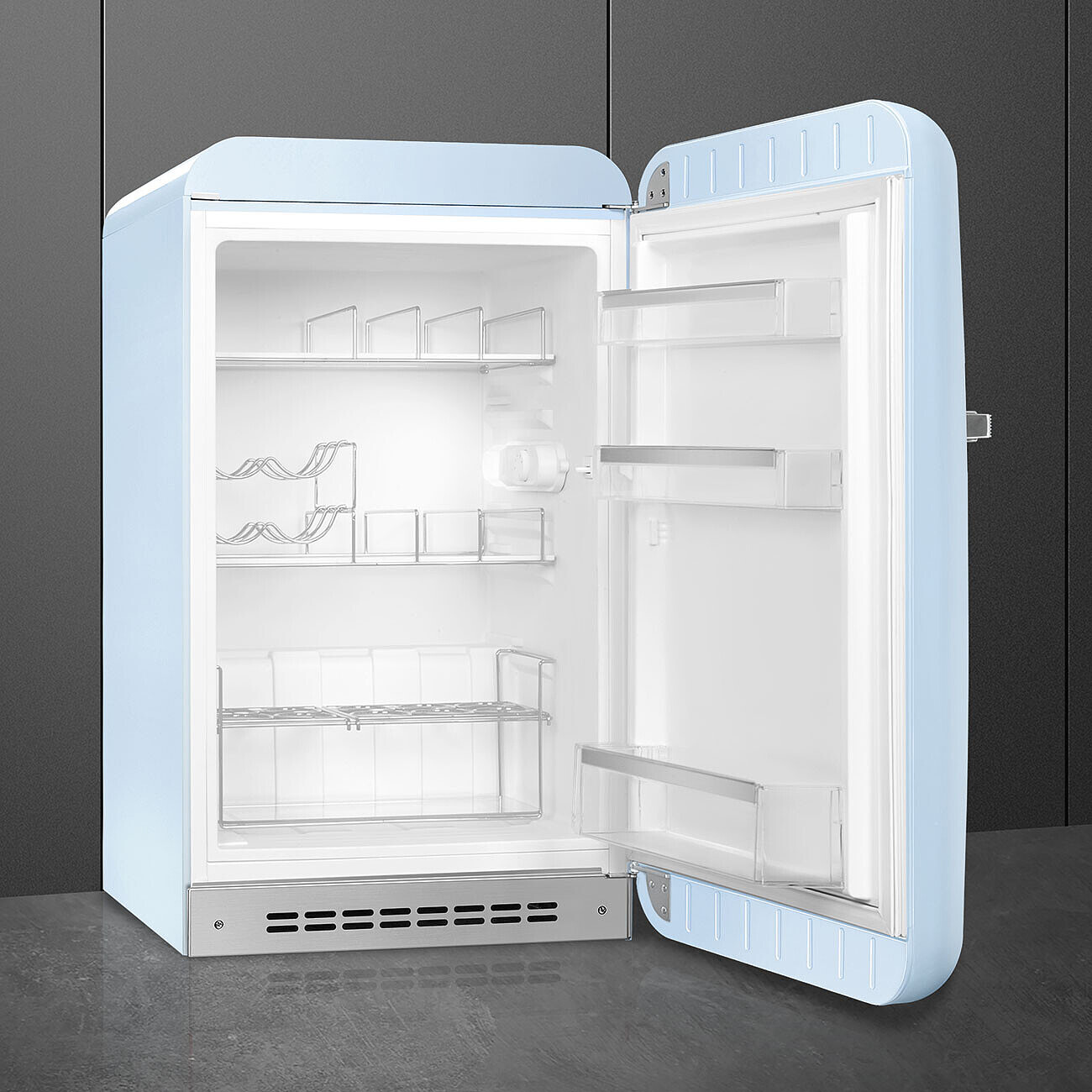 Un refrigerador retro azul claro con la puerta abierta. Se ven estantes interiores. El fondo es una pared y piso gris oscuro.