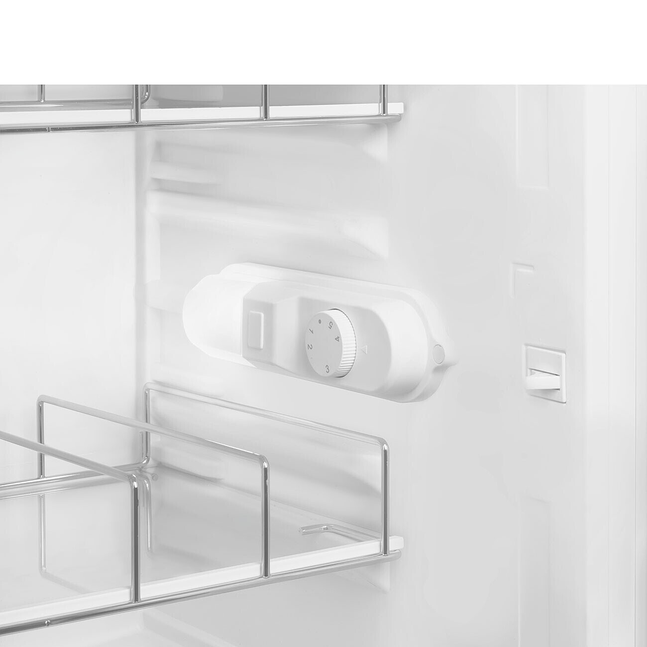 Dentro de un refrigerador blanco, se ve un dial de control de temperatura, junto con estantes de metal. El interior es mayormente blanco.