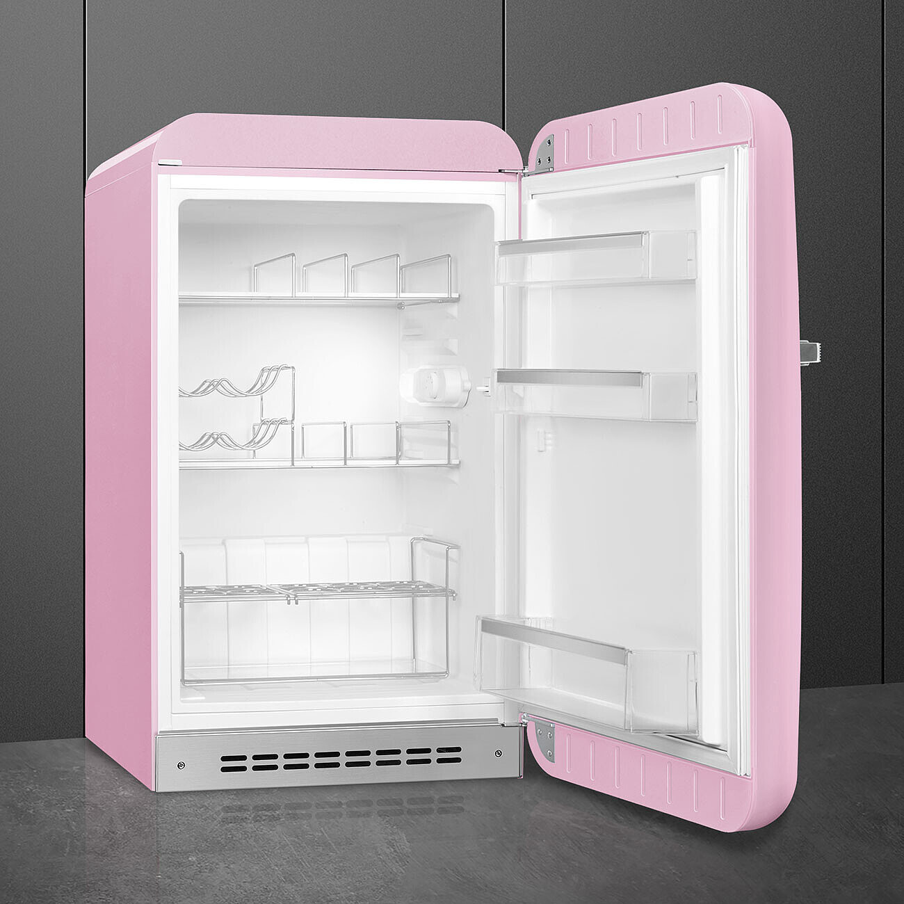 Un refrigerador rosa retro con la puerta abierta. Tiene estantes y compartimentos de almacenamiento. El fondo es una pared gris oscuro.