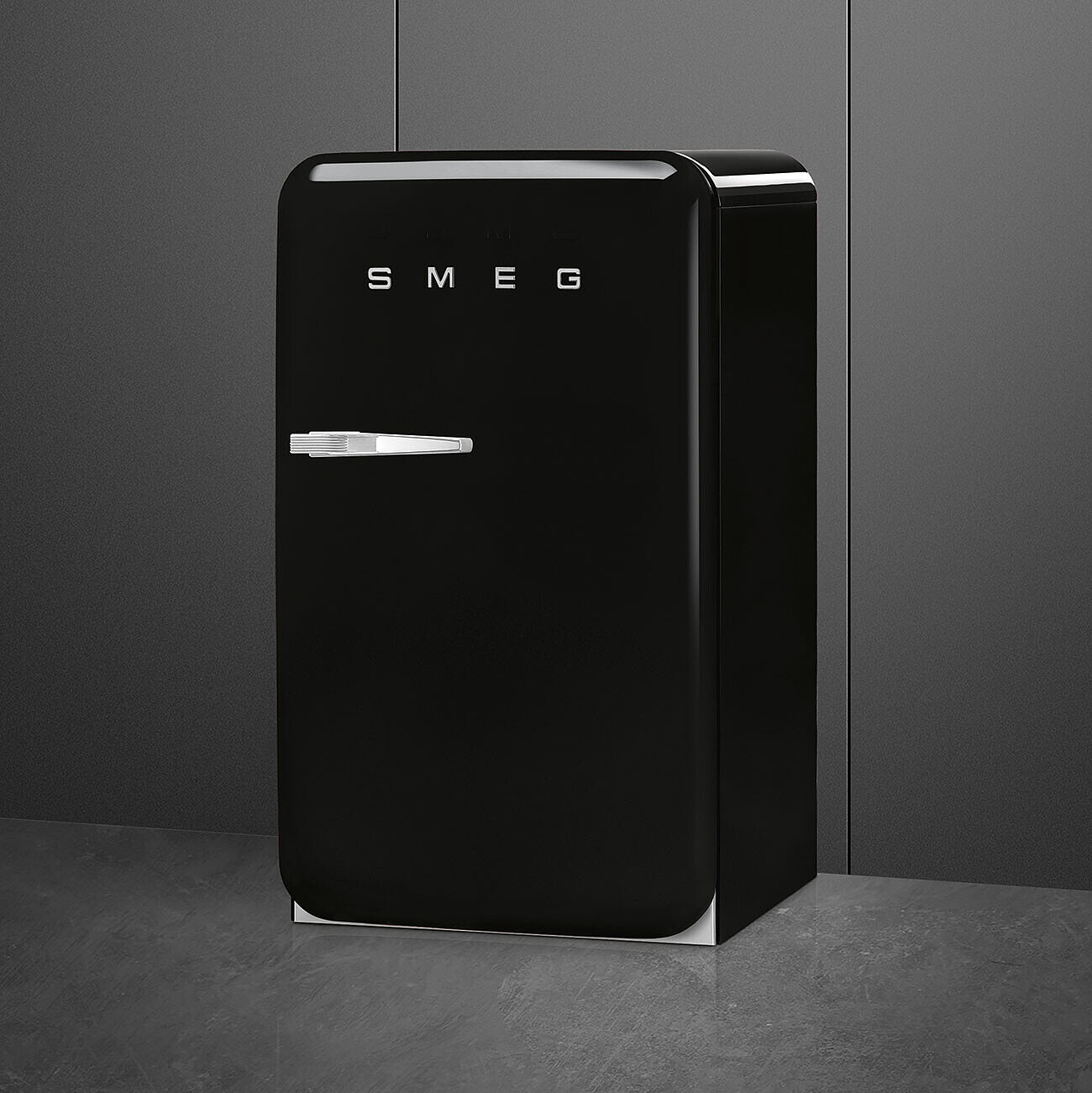 Un refrigerador negro SMEG se encuentra contra una pared gris. Tiene una manija y el logotipo de SMEG. El suelo es gris.