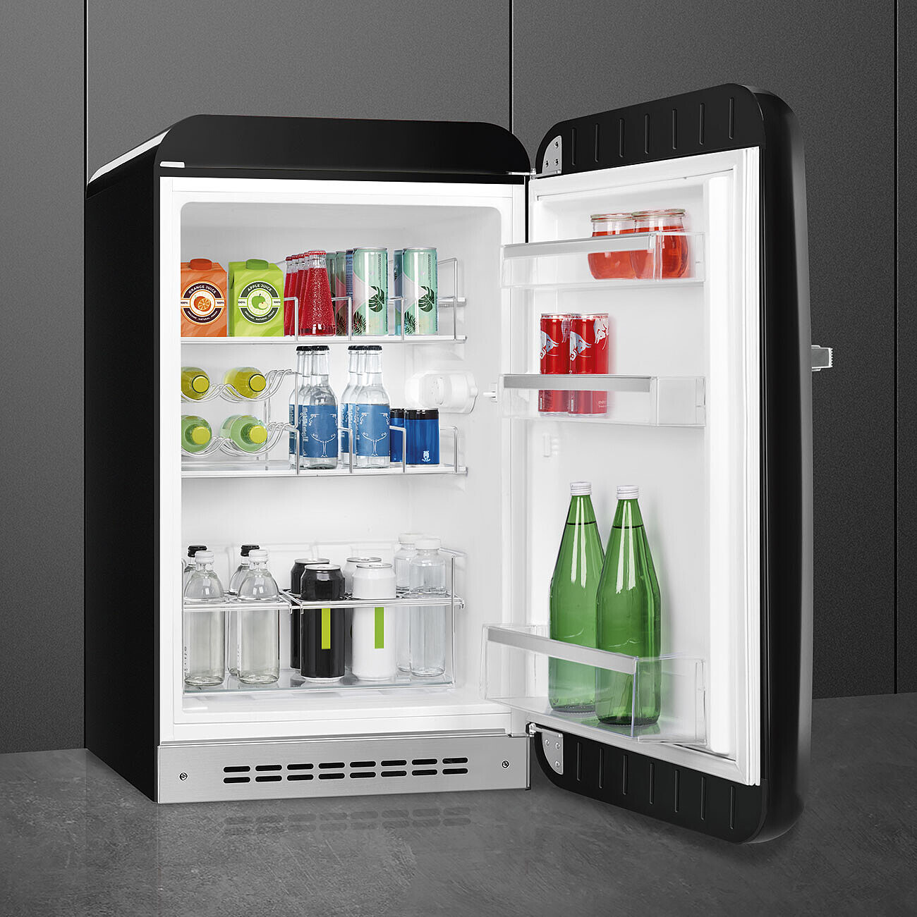 Un mini-refrigerador negro abierto, lleno de bebidas: cajas de jugo, latas de refresco, botellas de vidrio y frascos. La puerta está abierta, revelando el contenido.