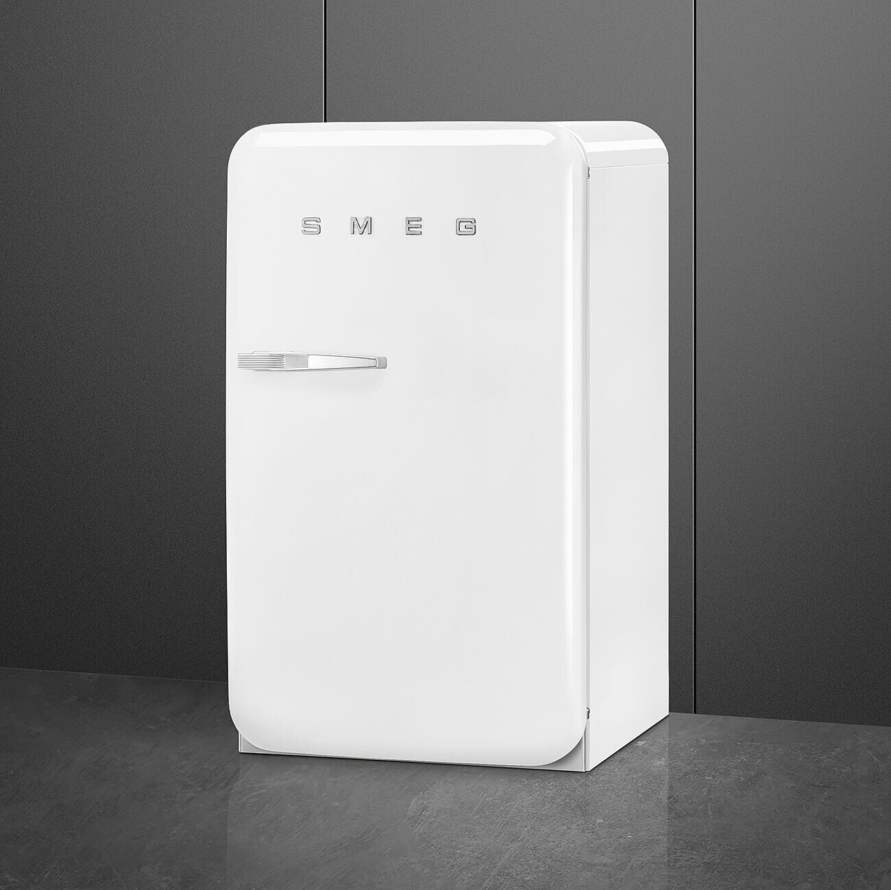 Un refrigerador Smeg blanco se encuentra contra una pared gris. Tiene una manija y el logo de Smeg. El suelo es gris oscuro.