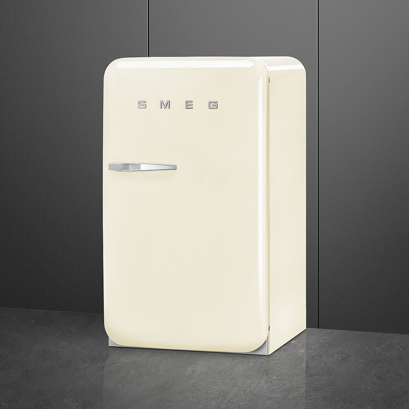 Un refrigerador SMEG crema de estilo retro sobre un suelo gris, contra una pared gris oscuro. Son visibles el tirador plateado y el logotipo SMEG.