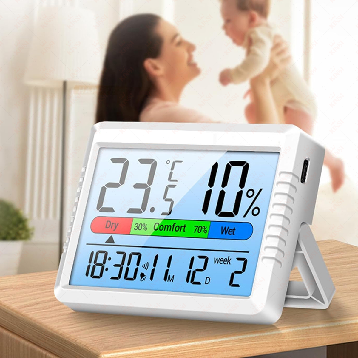 Ein digitales Thermometer zeigt 23,5°C Temperatur und 10% Luftfeuchtigkeit an. Im Hintergrund sind eine Frau und ein Baby unscharf.