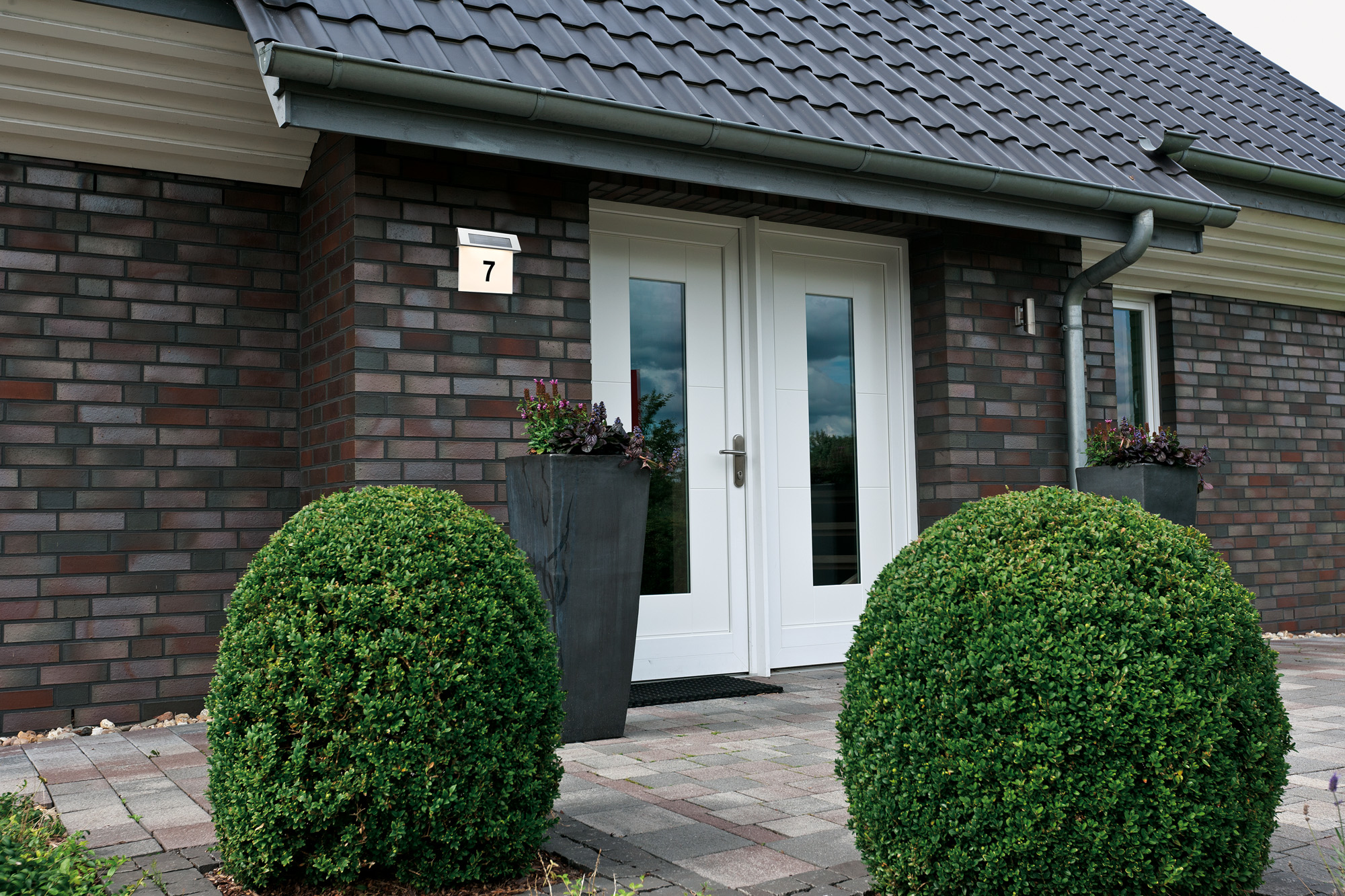 Wejście do domu z białymi podwójnymi drzwiami, ceglaną fasadą, zielonymi krzewami i znakiem z numerem 7. Widoczny jest również ciemny dach i doniczki.