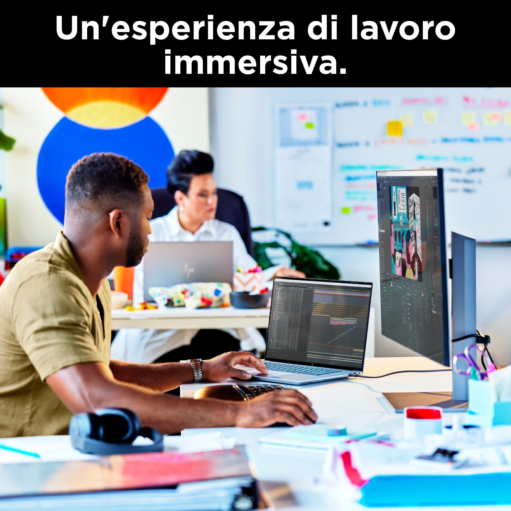 Un uomo lavora su un laptop a una scrivania. Un monitor grande mostra un'immagine. Un'altra persona lavora su un laptop sullo sfondo.