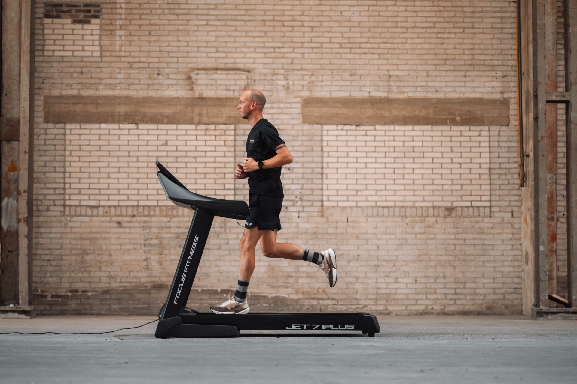 Man running on a treadmill indoors. He wears black shorts and shirt. The treadmill is black. Brick wall in background.
