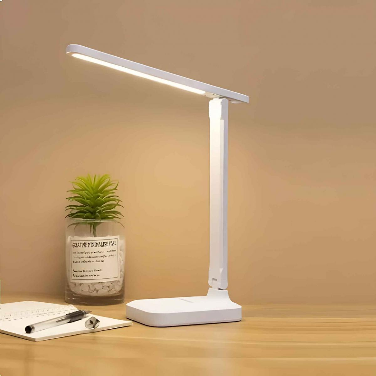 Eine weiße Schreibtischlampe steht auf einem Holzschreibtisch. Eine kleine Pflanze in einer Glasvase steht in der Nähe der Lampe. Ein Notizbuch und ein Stift sind ebenfalls sichtbar.