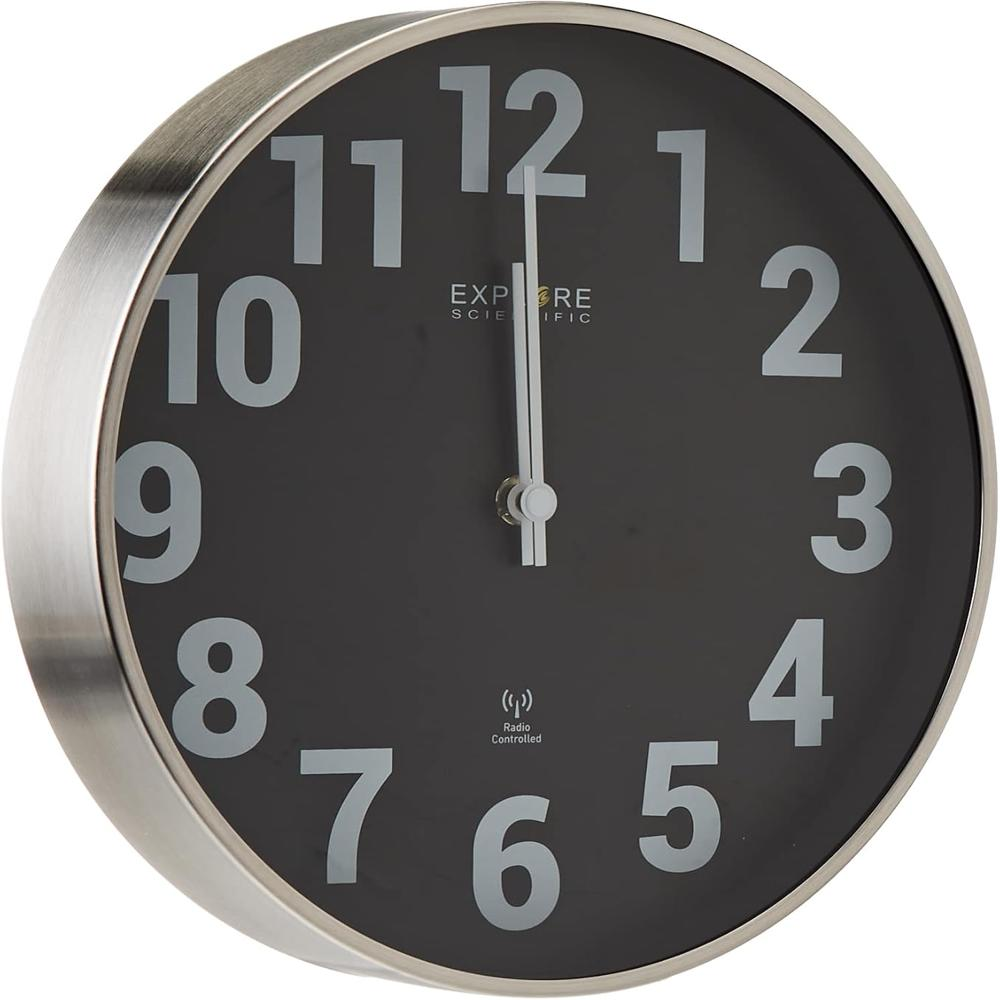 Un orologio rotondo con quadrante nero, numeri bianchi e cornice argentata. Le lancette puntano alle 12. Sono visibili 'Explore Scientific' e 'Radio Controlled'.