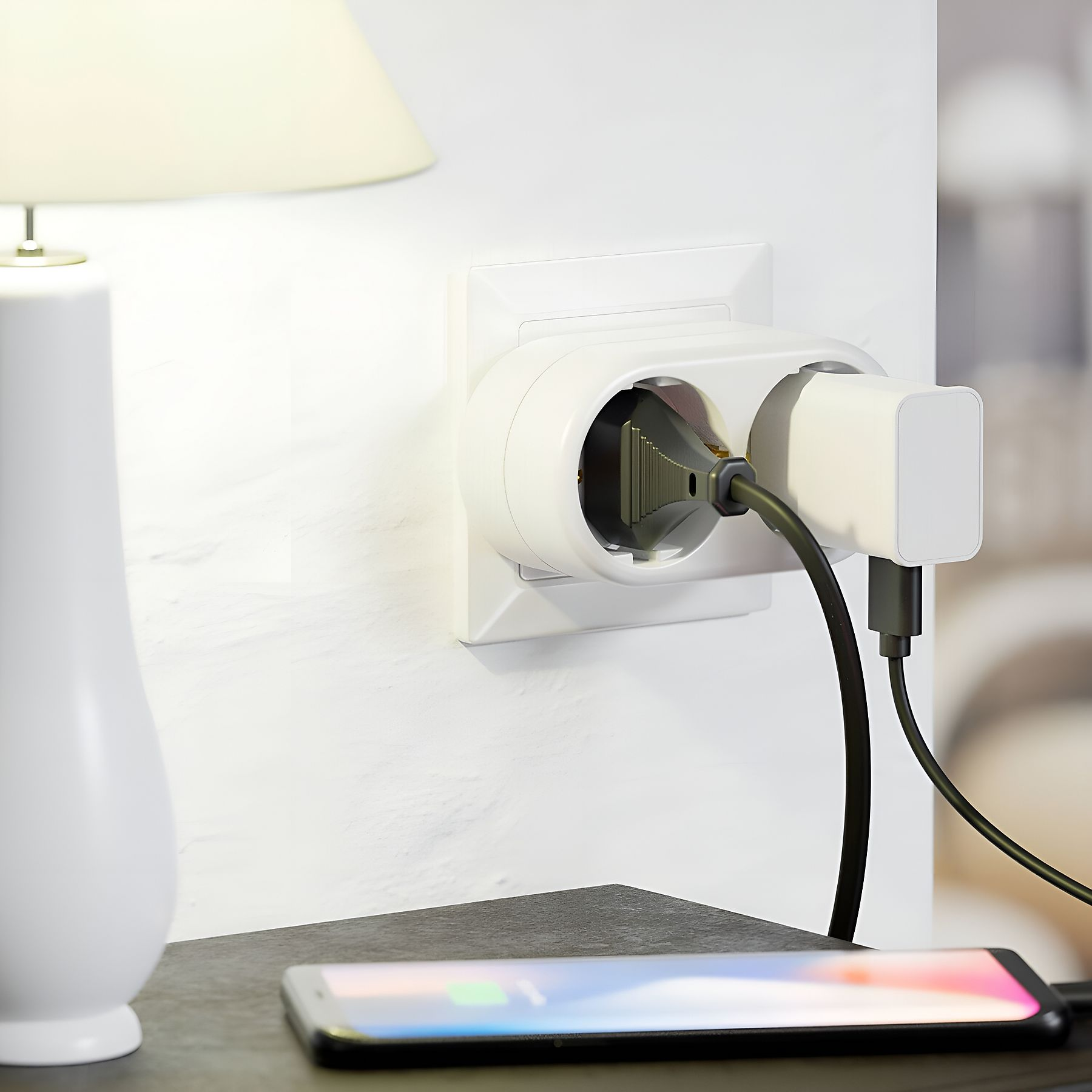 Eine weiße Wand mit Steckdose und Ladegerät. Ein Telefon liegt unten und lädt. Links ist eine Lampe.