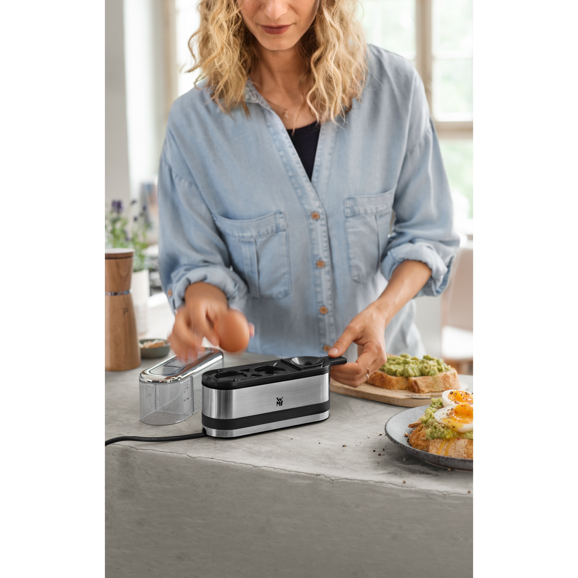 Kobieta wbija jajko do jajowaru. Tosty z awokado i jajkiem na talerzu. Ustawienia kuchenne.