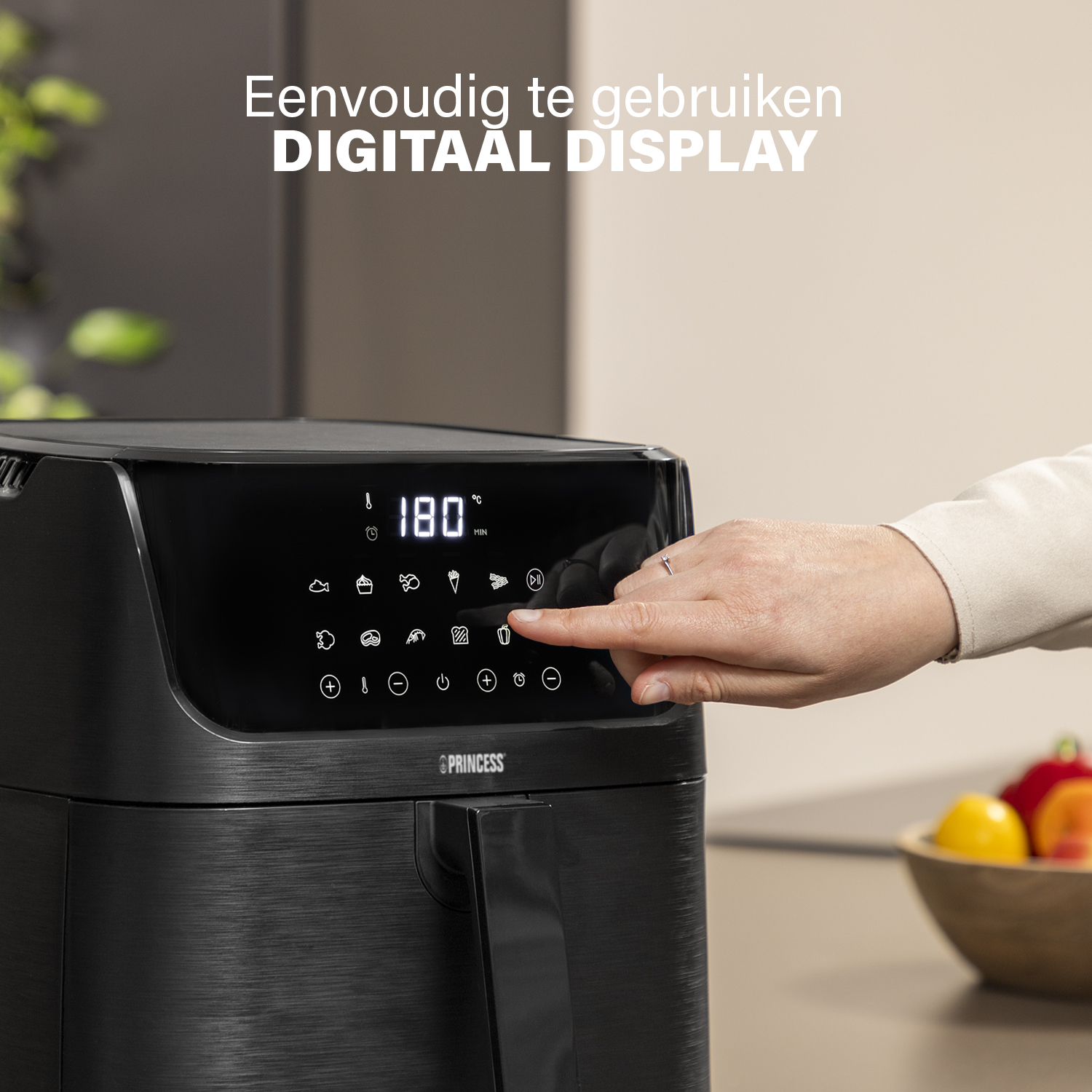 Ein schwarzer Airfryer mit Digitalanzeige. Eine Hand berührt den Bildschirm. Früchte in einer Schüssel im Hintergrund.