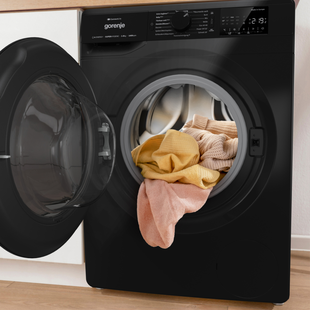 Eine schwarze Waschmaschine mit offener Tür, die bunte Kleidung im Inneren zeigt. Die Maschine steht auf einem Holzboden.
