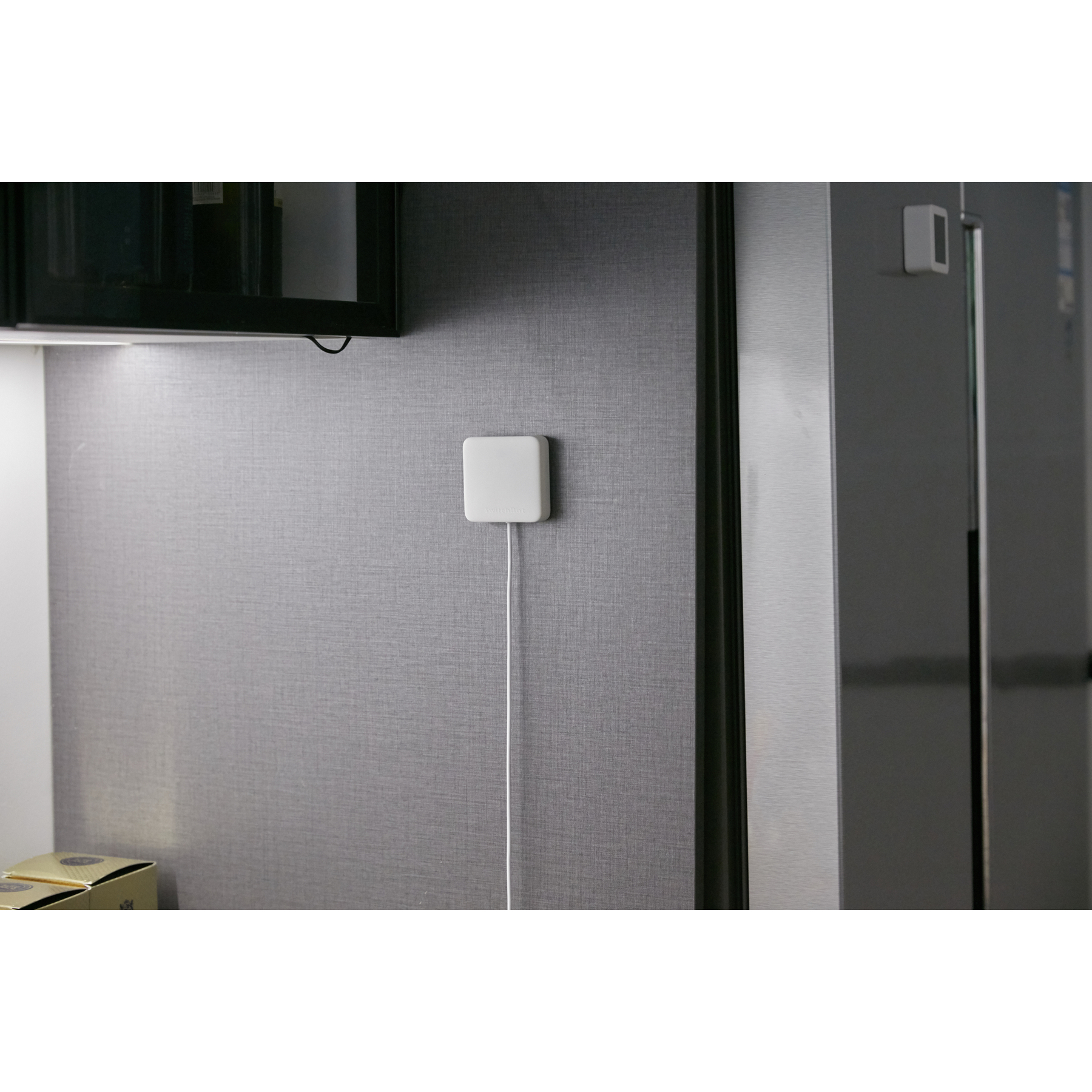 Ein weißer Sensor ist an einer grauen Wand montiert. Ein Kabel erstreckt sich nach unten. Über und rechts befindet sich ein schwarzer Schrank.