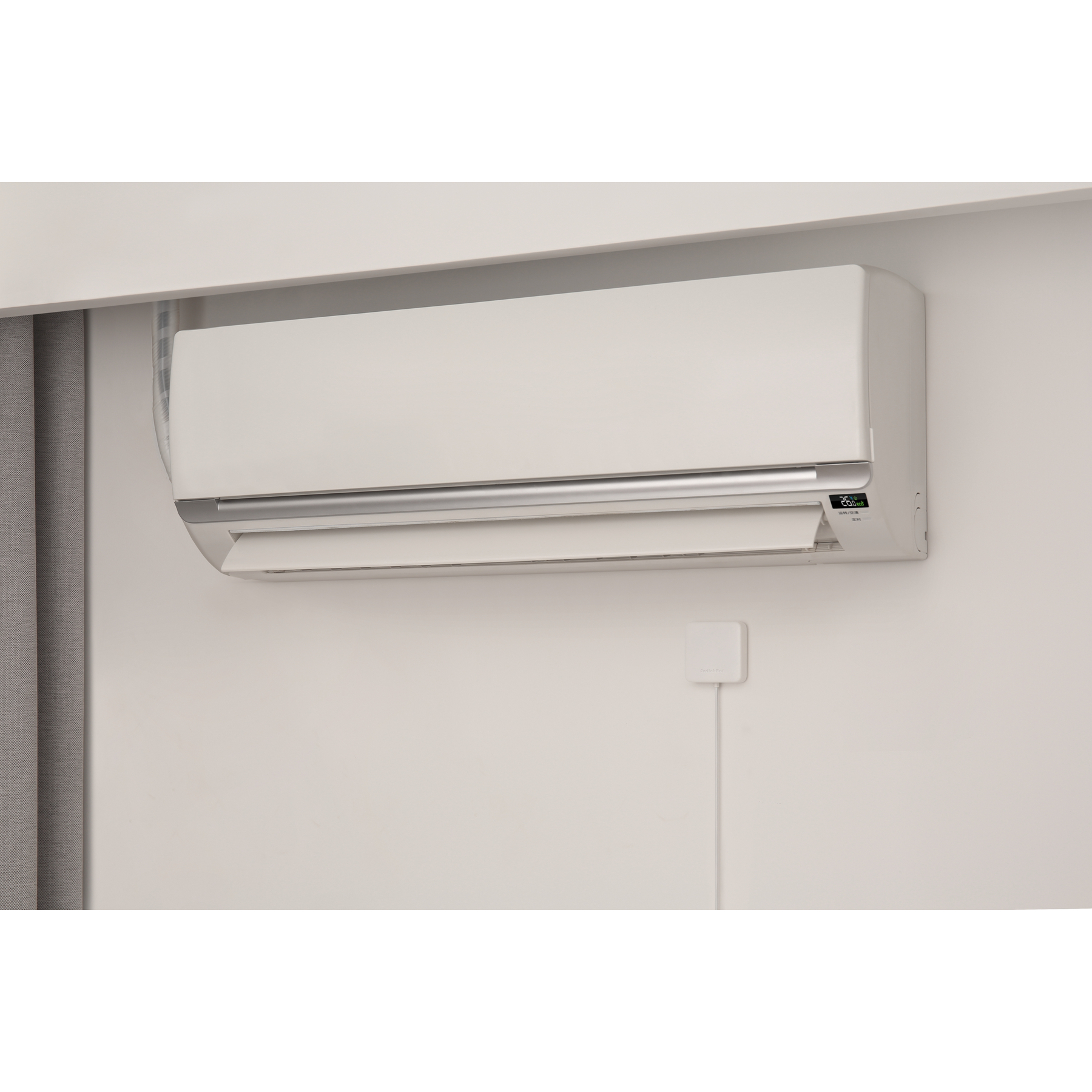 Weißer Klimaanlage an einer hellen Wand. Ein kleines Bedienfeld befindet sich darunter mit einem Kabel. Grauer Vorhang links.