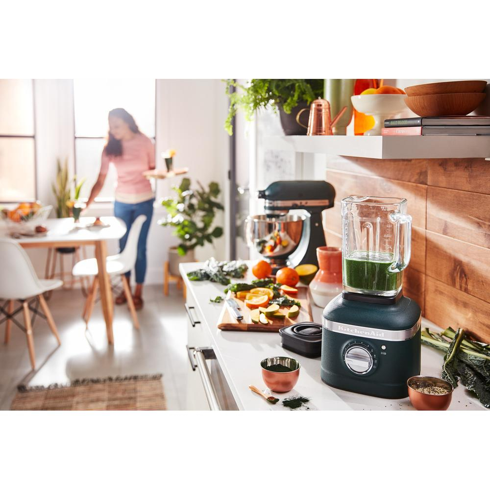 Küchenszene: ein Mixer mit grünem Smoothie, Früchten, Gemüse und eine Frau, die Essen zubereitet. Verschiedene Küchengeräte und Zutaten.