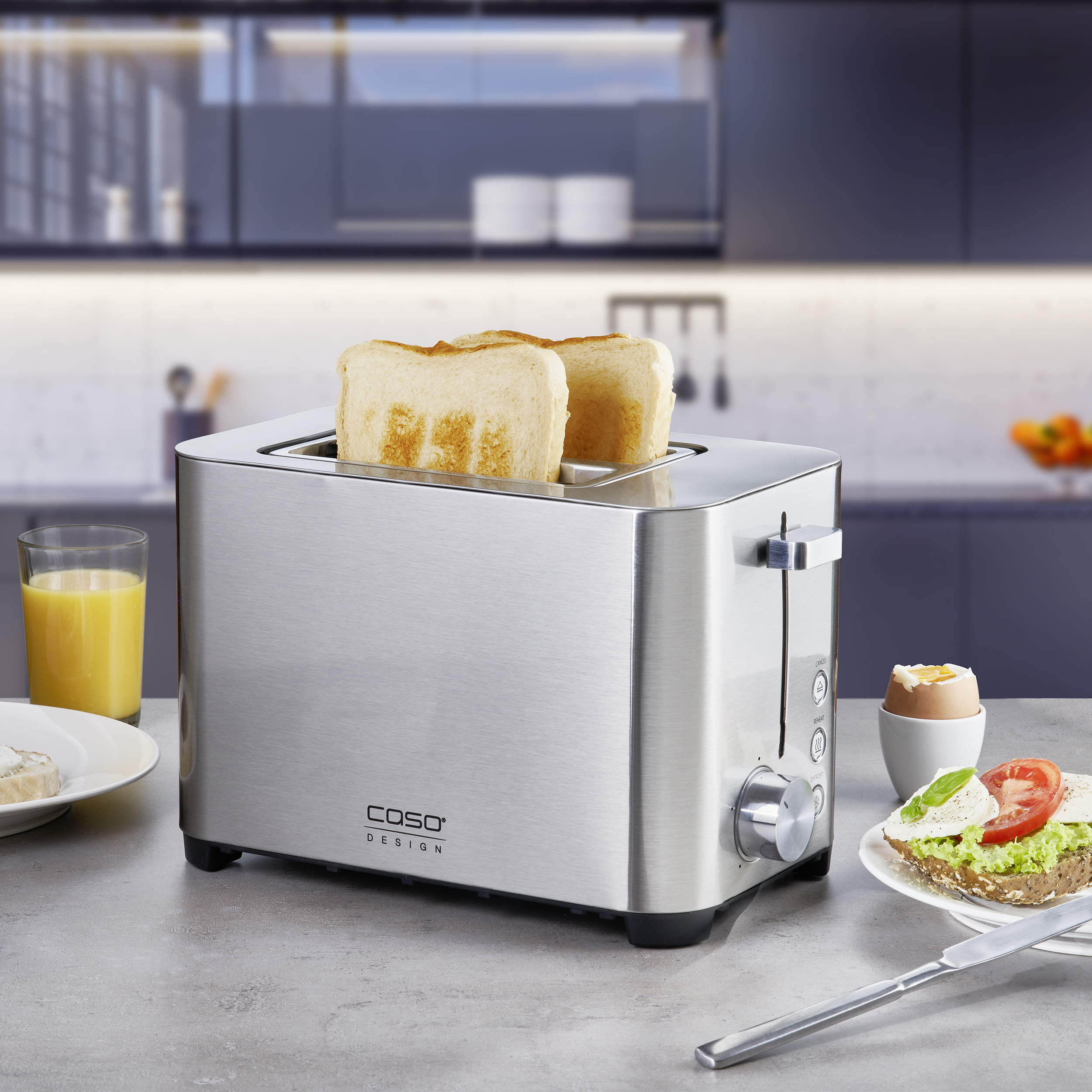 Ein Edelstahl-Toaster mit zwei Toastscheiben. Ebenfalls vorhanden: Saft, Ei und ein Sandwich auf einem Tisch.