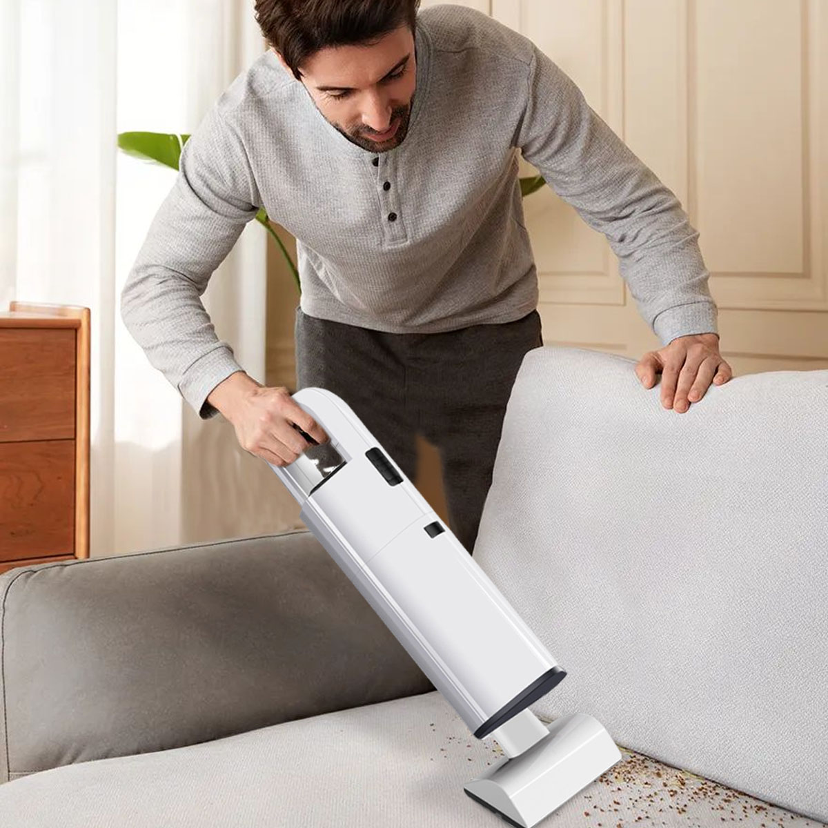 Ein Mann saugt mit einem Handstaubsauger ein Sofa ab. Das Sofa ist grau, und der Staubsauger ist weiß. Der Mann trägt ein graues Hemd.
