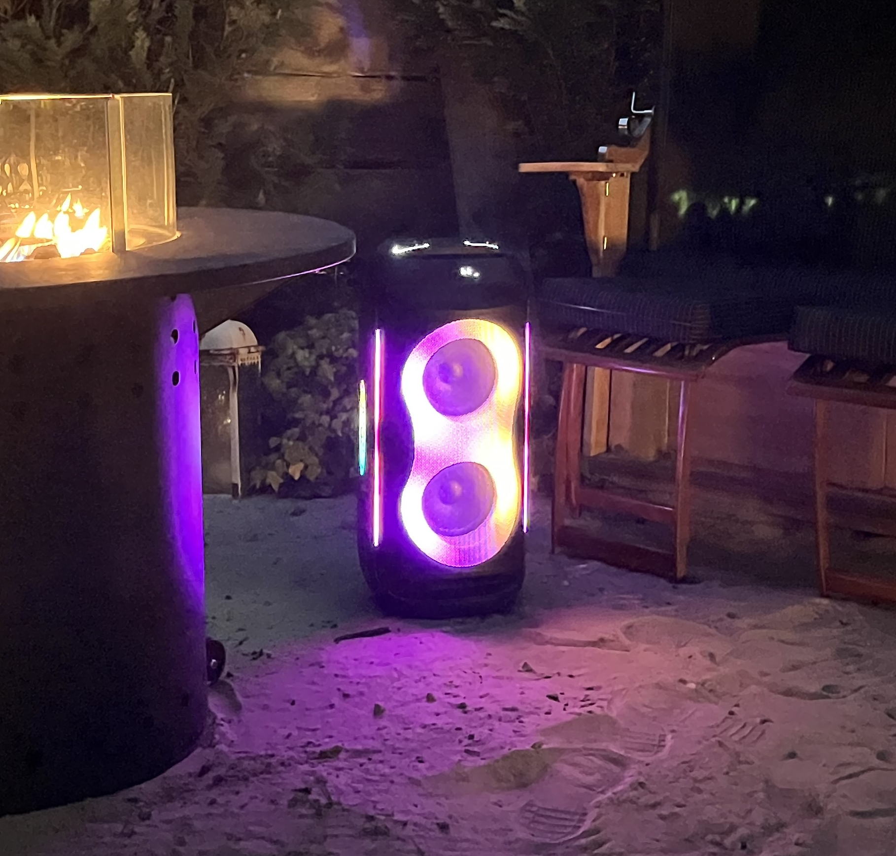 Ein beleuchteter Lautsprecher steht auf Sand, mit einer Feuerstelle und Holzmöbeln in der Nähe. Der Lautsprecher leuchtet lila und orange.