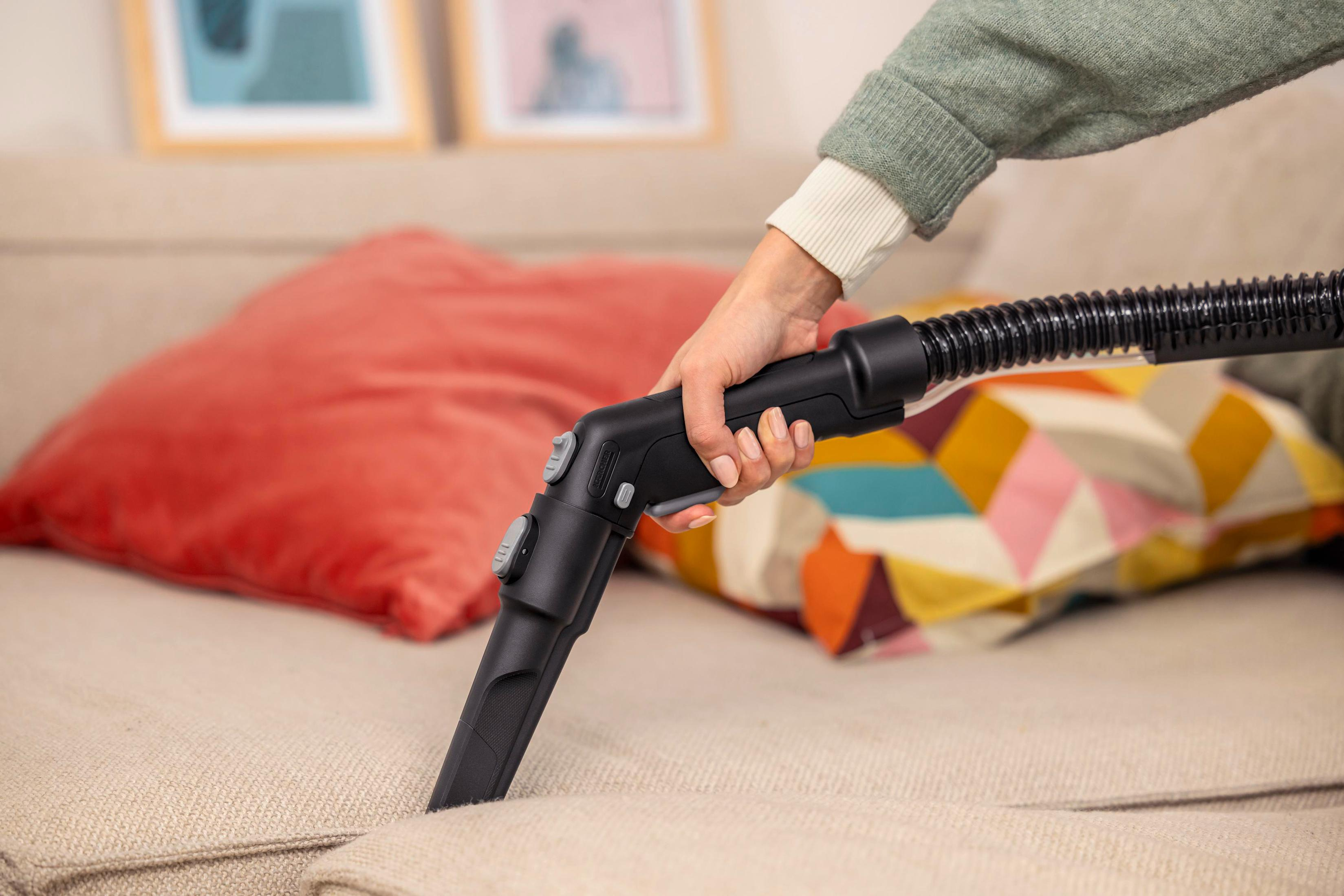 Eine Person saugt ein Sofa ab. Ein schwarzer Staubsaugerkopf befindet sich auf dem beigefarbenen Stoff. Ein rotes Kissen und bunte Kissen sind im Hintergrund.