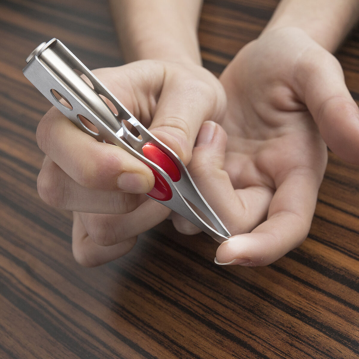 Una persona tiene delle pinzette con un accento rosso. Lo sfondo è un tavolo di legno.