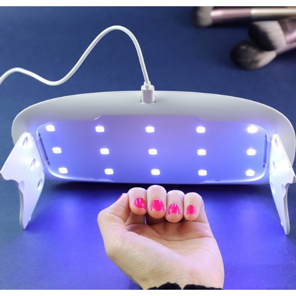 Eine Hand mit pinken Nägeln unter einer UV-Lampe. Die Lampe ist weiß, mit blauem Licht. Im Hintergrund sind Make-up-Pinsel.