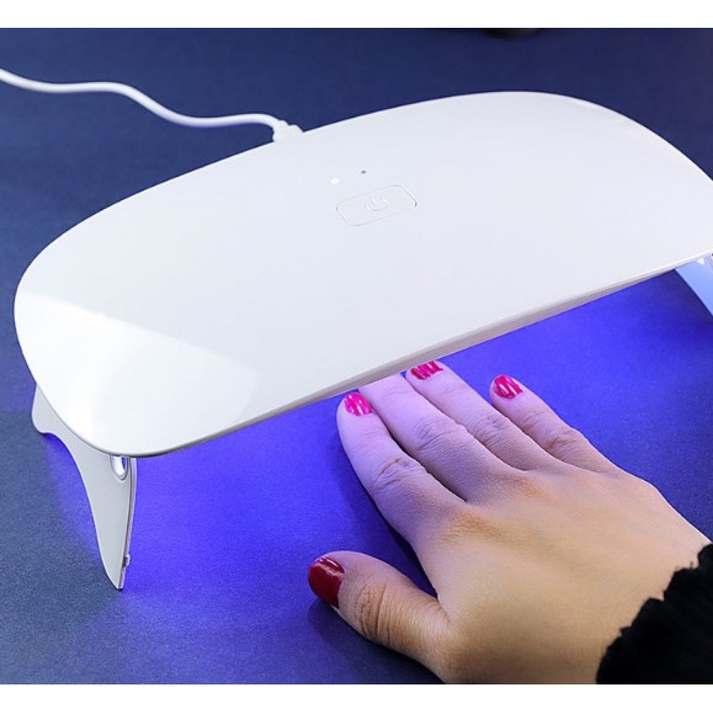 Eine Hand mit rotem Nagellack unter einer weißen UV-Lampe. Die Lampe steht auf einer blauen Oberfläche. Die Szene deutet auf eine Maniküre hin.