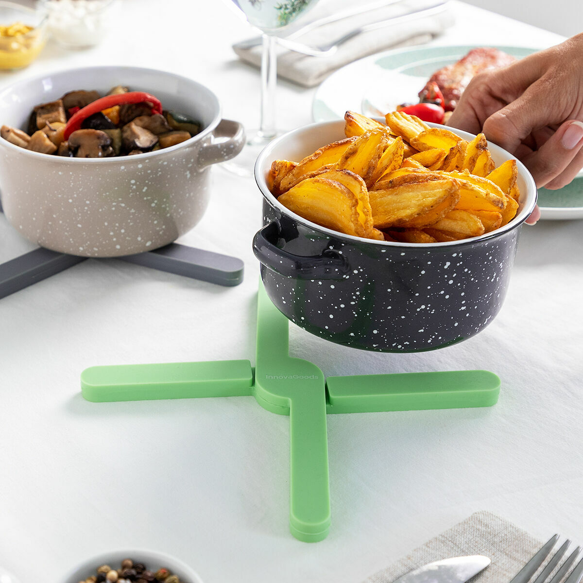 Zwei gesprenkelte Töpfe auf Untersetzern halten Essen. Einer hat Pommes, der andere Gemüse. Eine Hand hält den Pommes-Topf. Weiße Tischdecke.