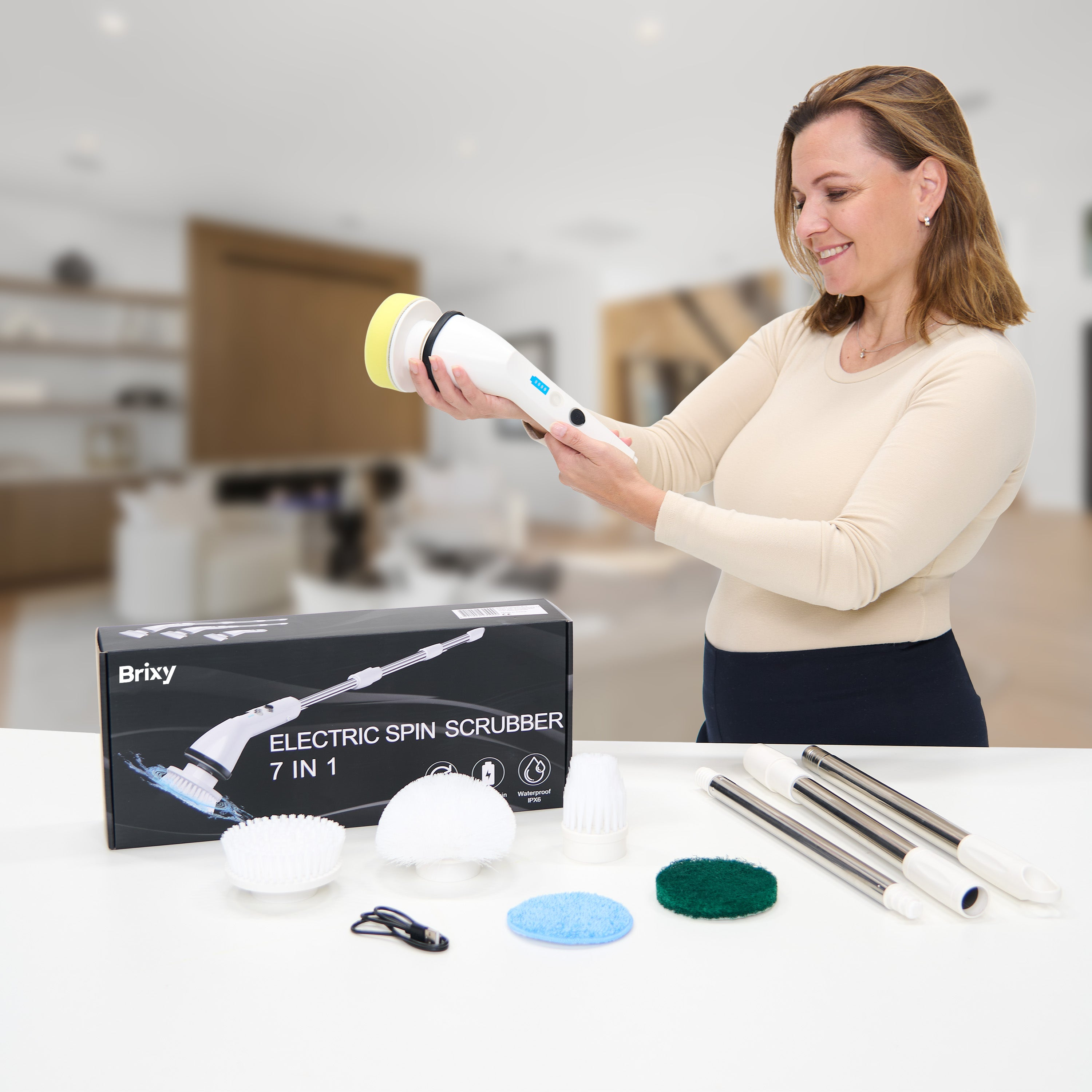 Woman holding a white and yellow electric spin scrubber. Box and scrubber heads are on a white surface. Blurred background.